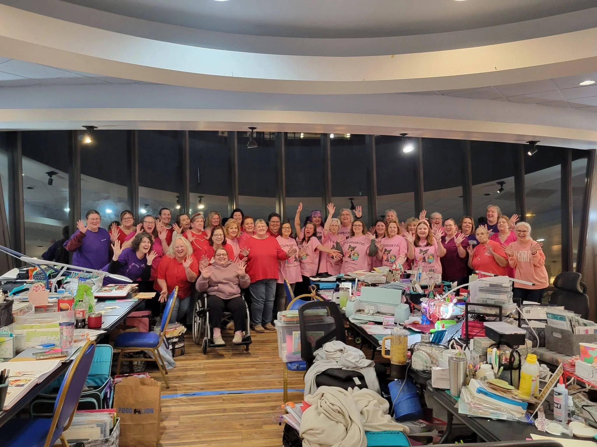 A large group of women, some in wheelchairs, smiling and waving at the camera inside a busy craft or sewing room with tables cluttered with craft supplies, fabrics, and tools.