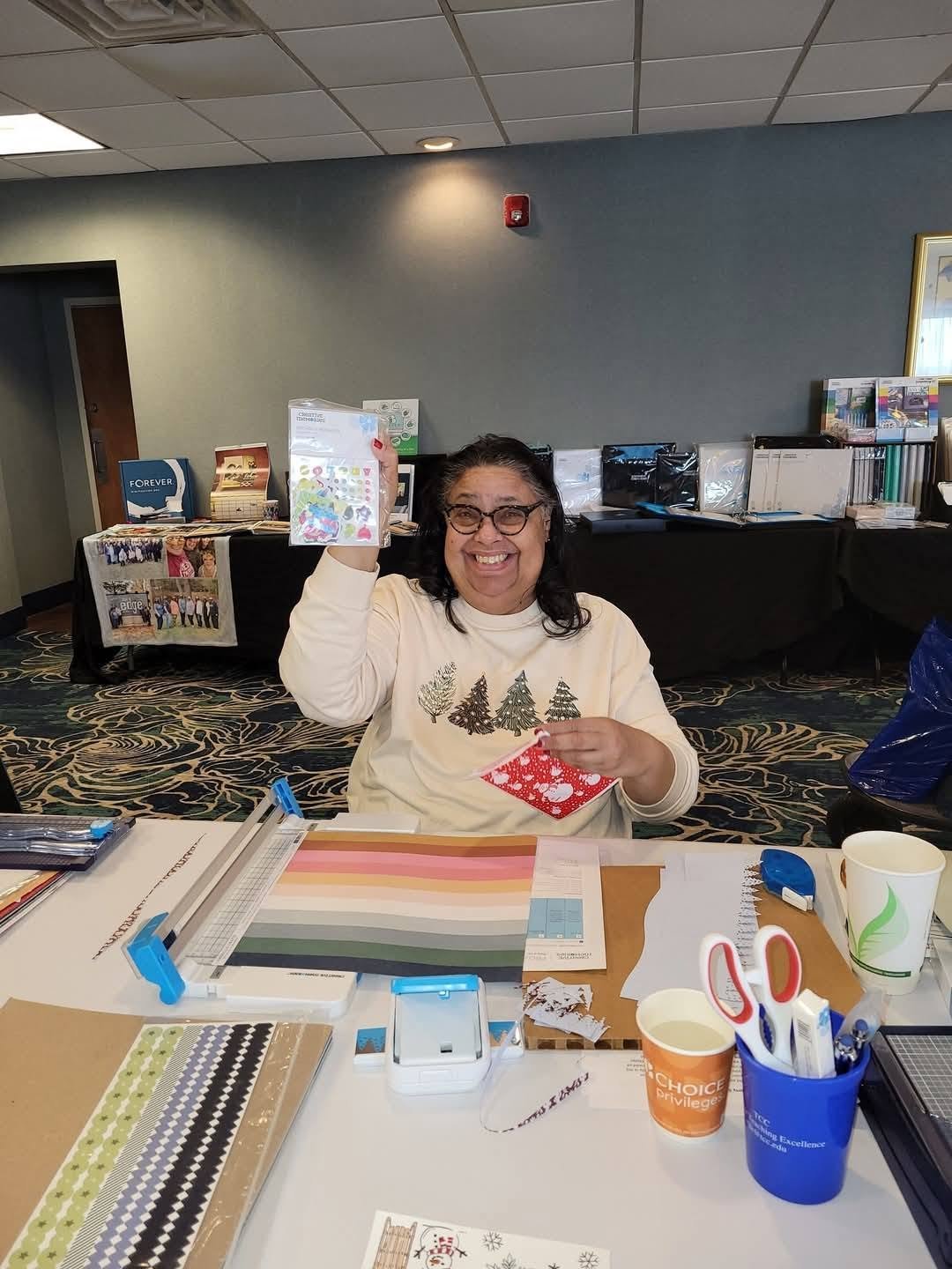 A woman smiling and holding up a small holiday-themed sticker sheet in a craft or office supplies room, with various stationery and craft materials on the table in front of her.