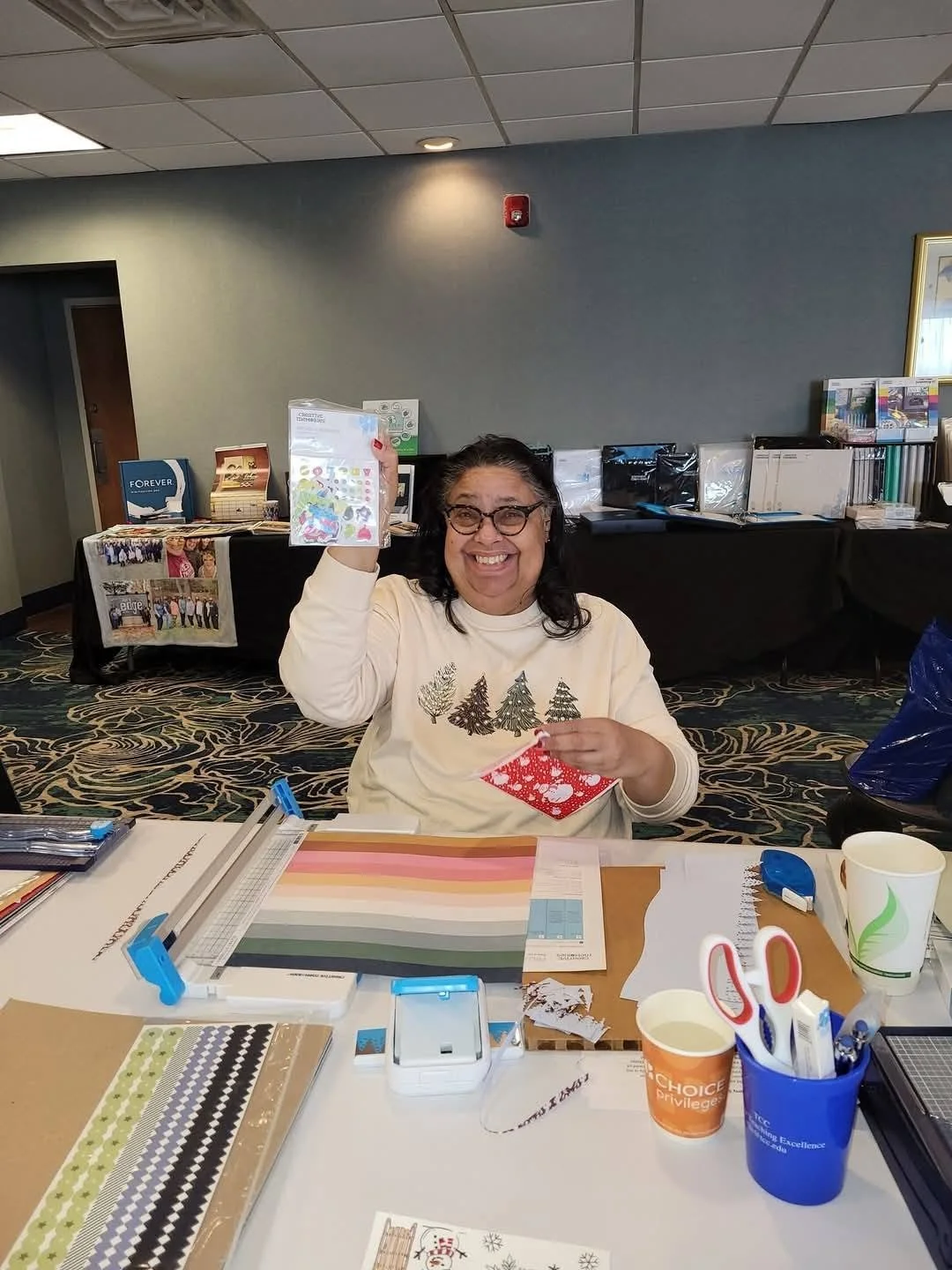 A woman smiling and holding a Christmas-themed craft project, seated at a table with crafting supplies in a well-lit room.