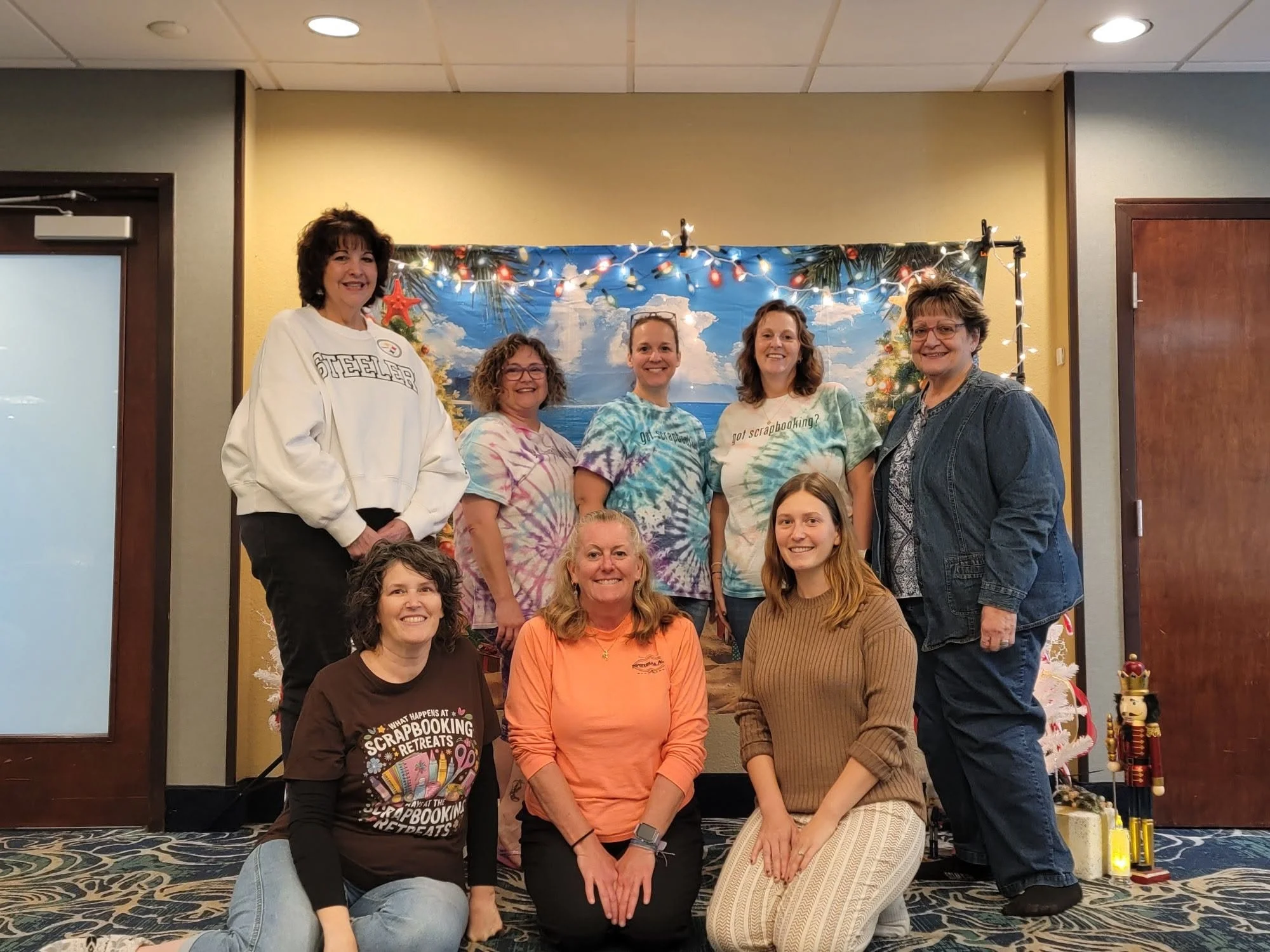 A group of ten women gathered together in a room decorated for Christmas, with a painted backdrop of a beach scene and Christmas lights. They are smiling and posing for the photo.