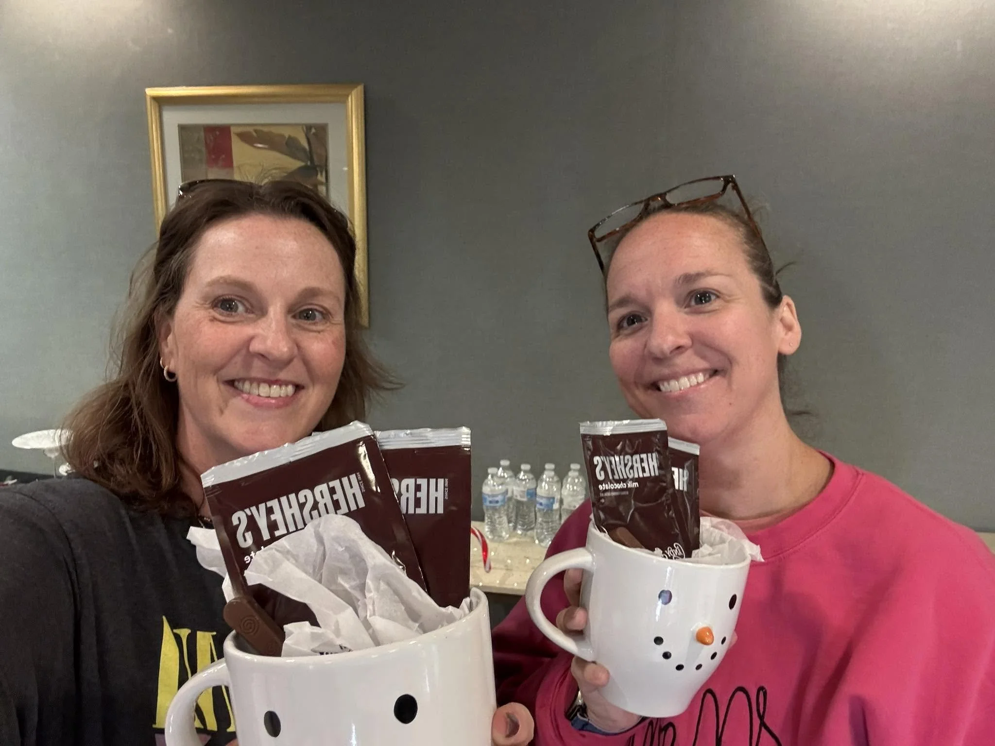 Two women smiling and holding snowman-shaped mugs with hot beverages, in an indoor setting with a gray wall and a framed artwork in the background.