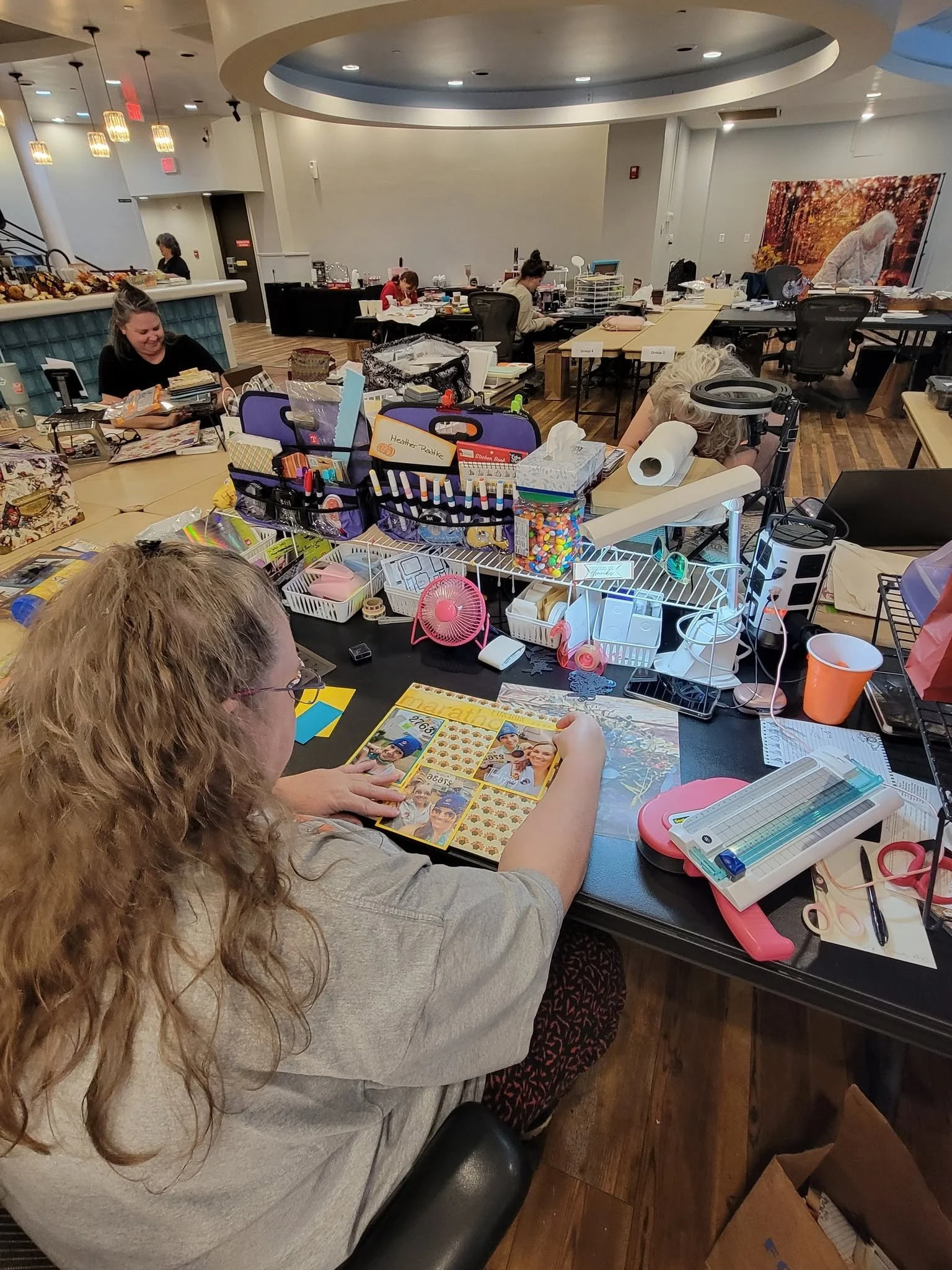 Multiple women are crafting at a large table cluttered with various art supplies, papers, and decorations in a well-lit room with wood floors.