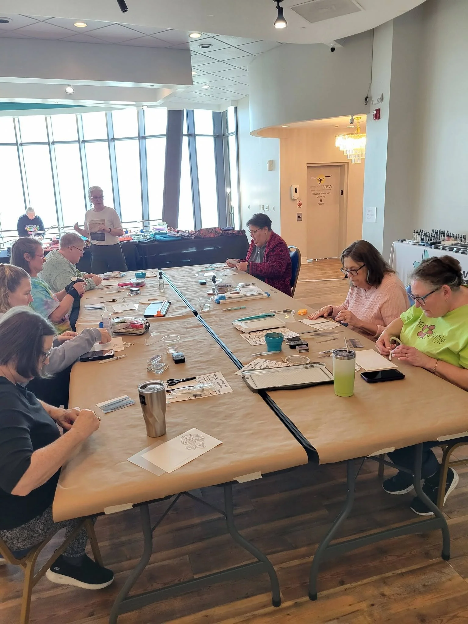 Group of women seated around a large table engaging in arts and crafts, with various supplies, in a bright, modern space with large windows.