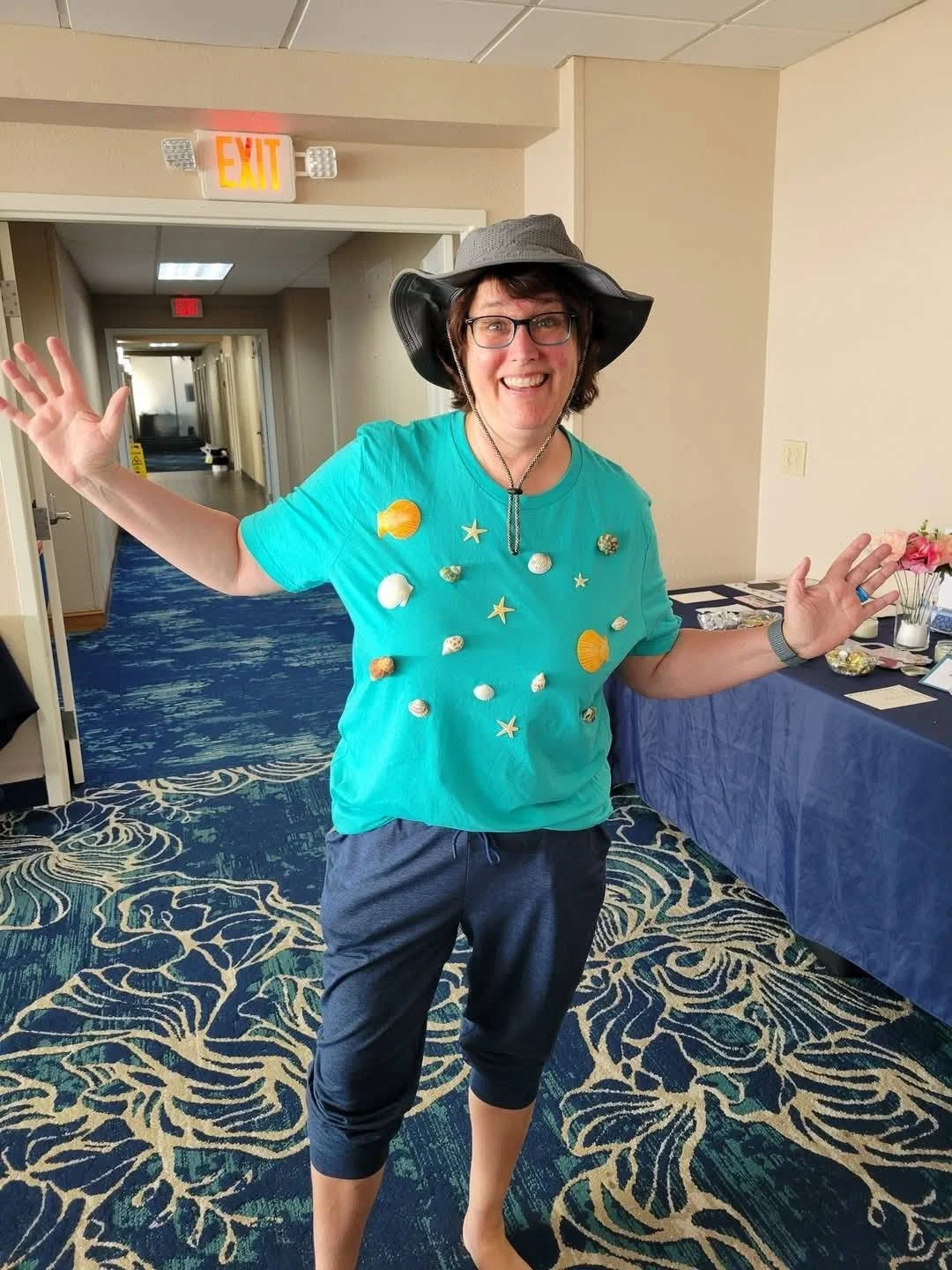 Woman smiling and waving, wearing a turquoise shirt decorated with shells and starfish, black pants, and a wide-brimmed hat, standing in a corridor with blue patterned carpet.