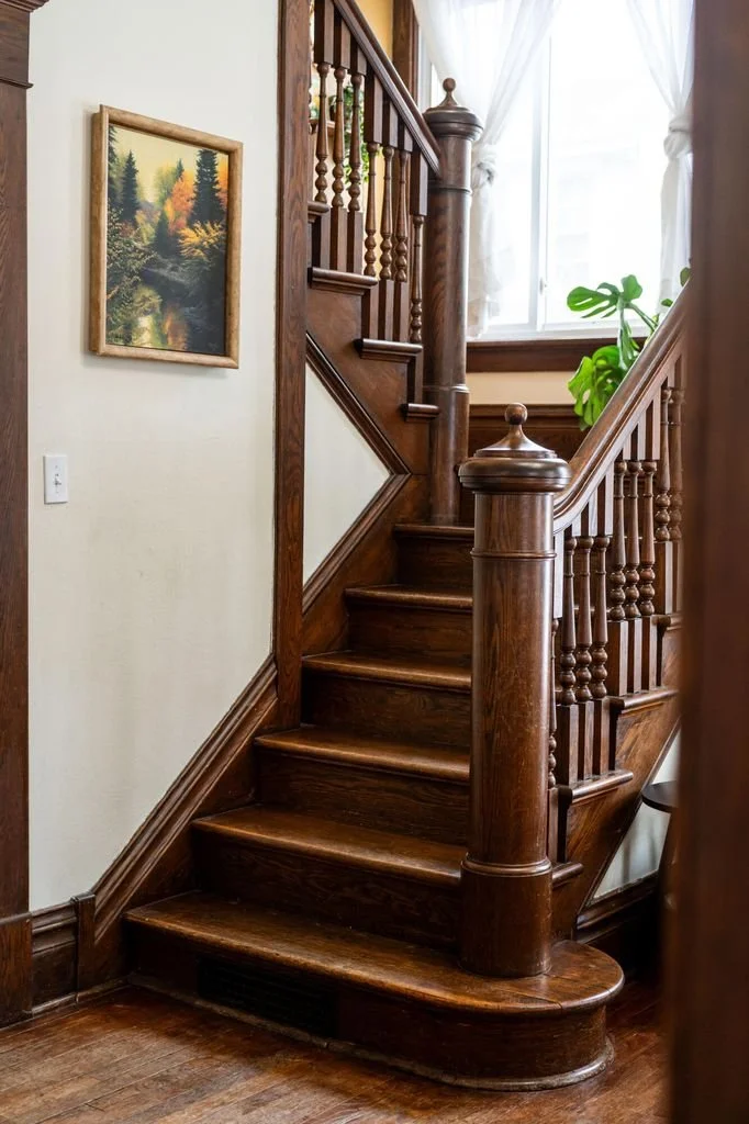 A wooden staircase with polished steps, carved newel posts, and spindles, located in a well-lit interior with white walls and a window with white curtains.