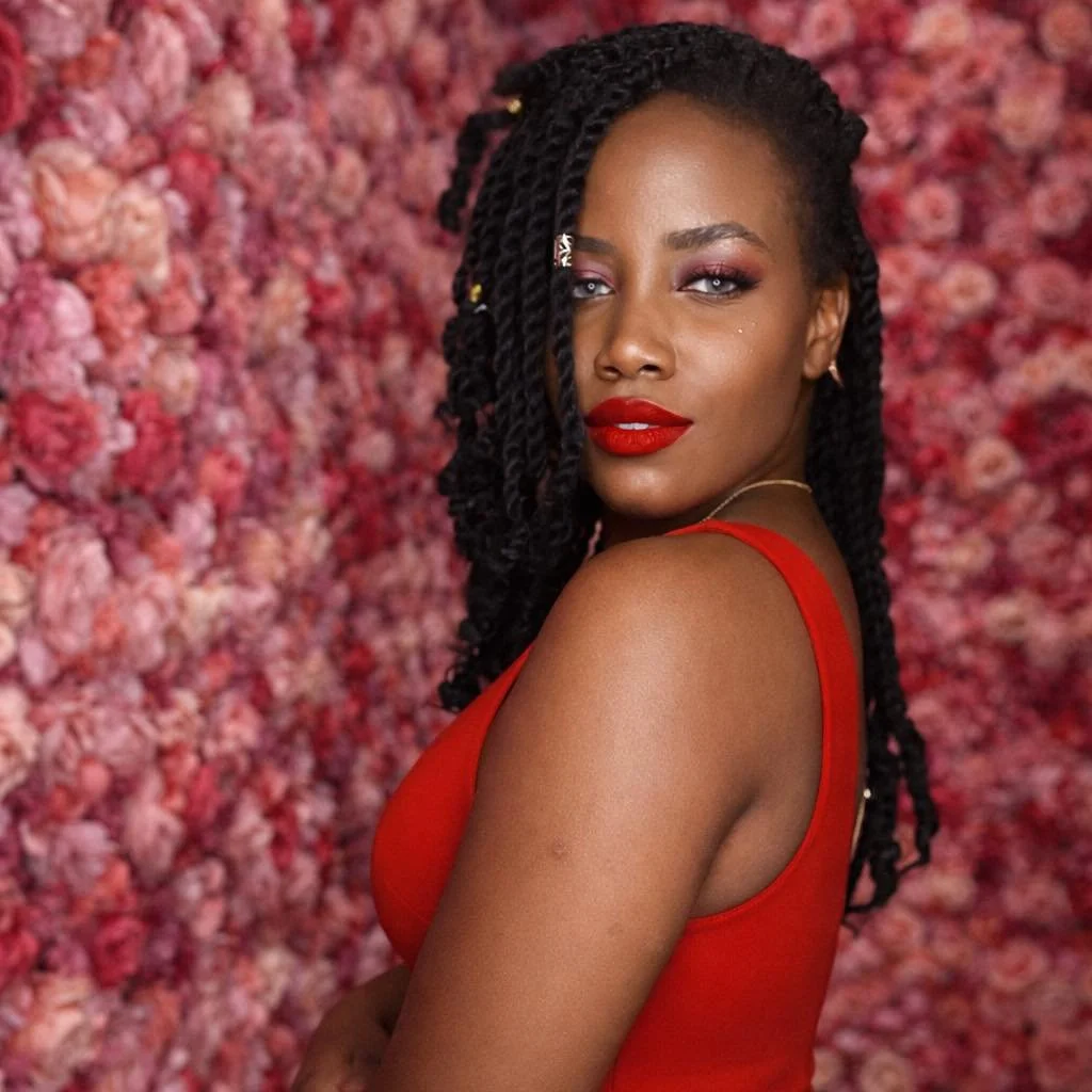 A woman with dark skin wearing a red dress and red lipstick, with long twisted hair and a confident expression, standing in front of a pink floral background.