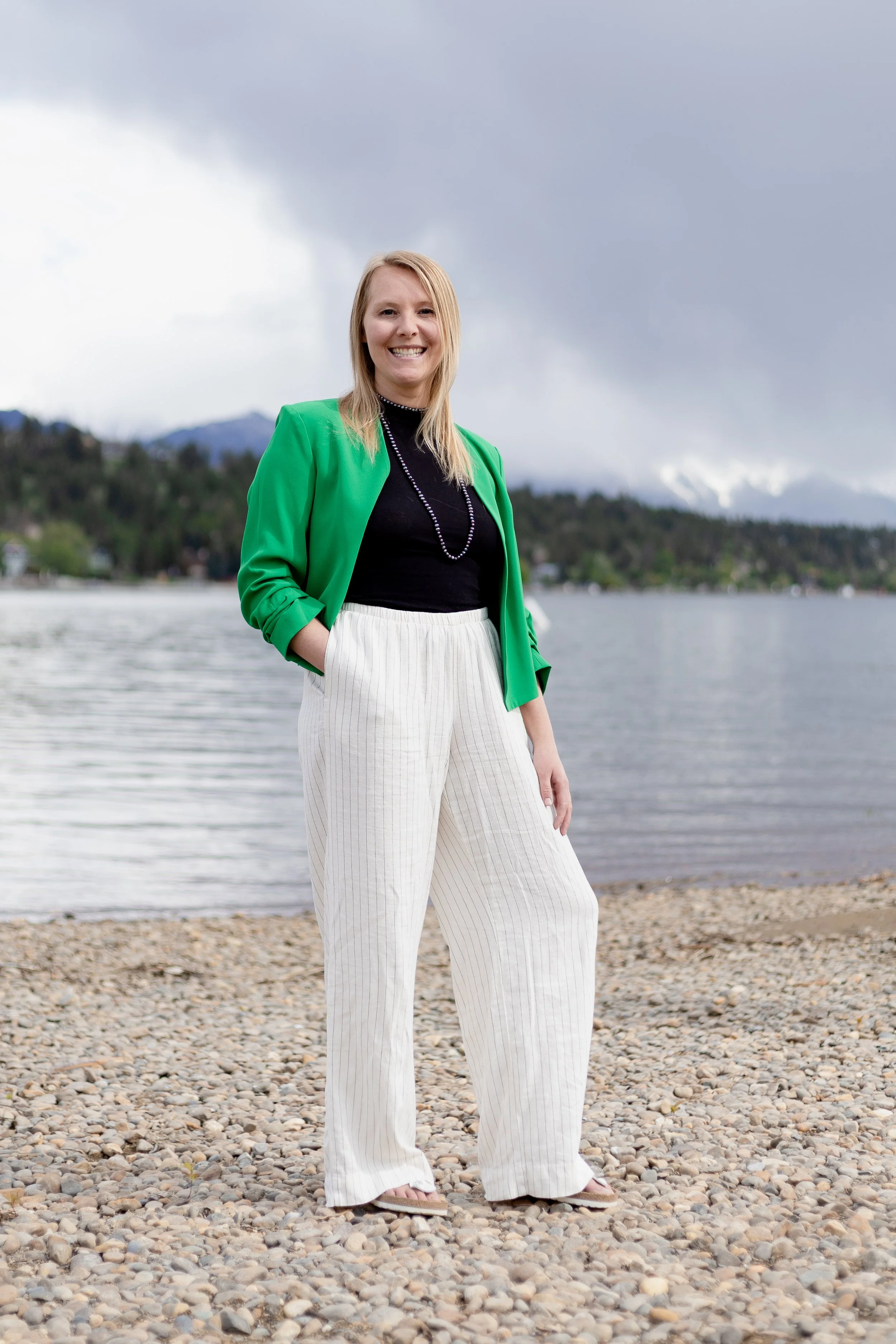 A woman standing on a rocky beach near water with mountains and cloudy sky in the background, smiling at the camera.