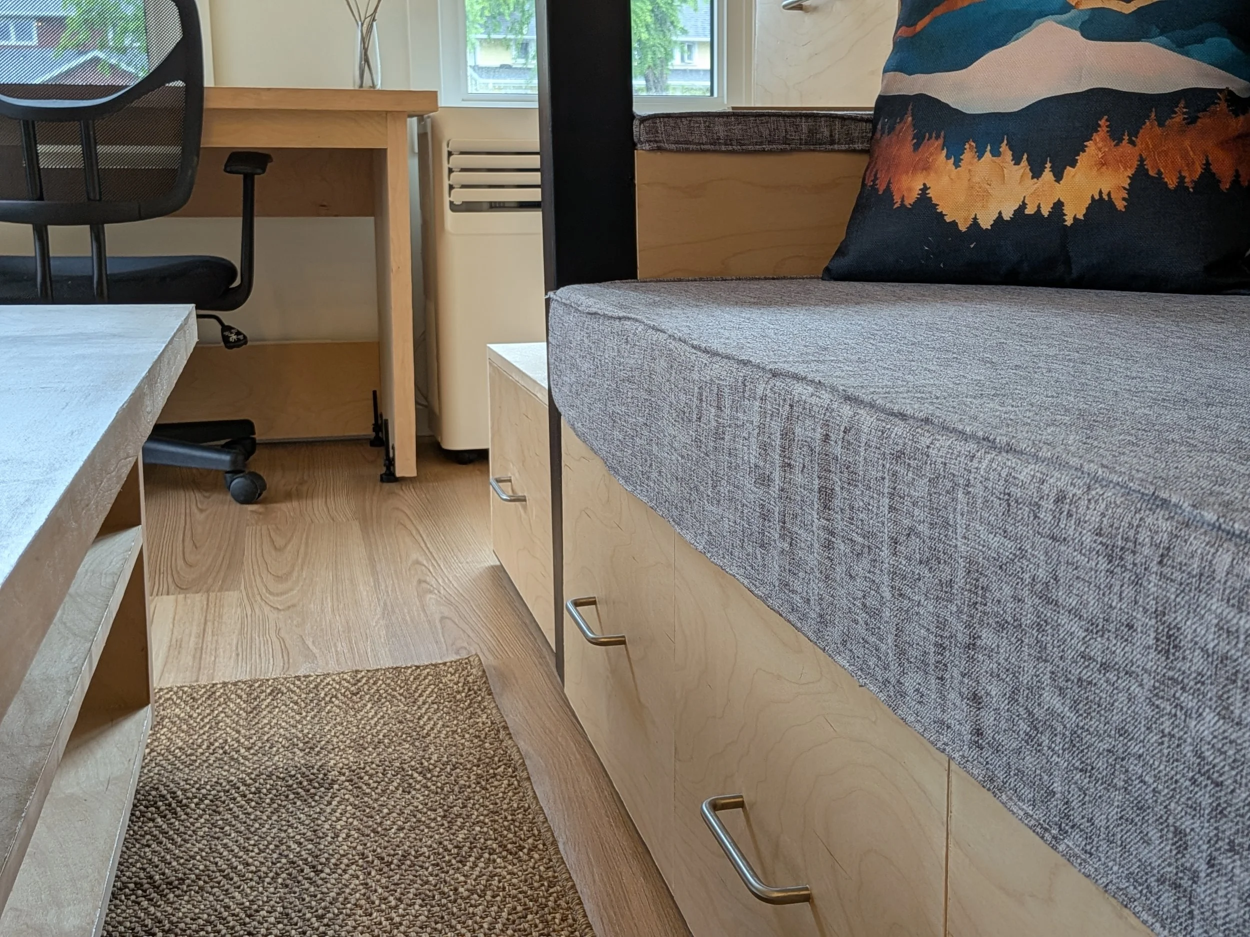 Interior of a living room or office with a gray fabric couch, a throw pillow with a mountain and forest scene, a wooden desk, a black office chair, and a window with a view of trees and neighboring buildings.
