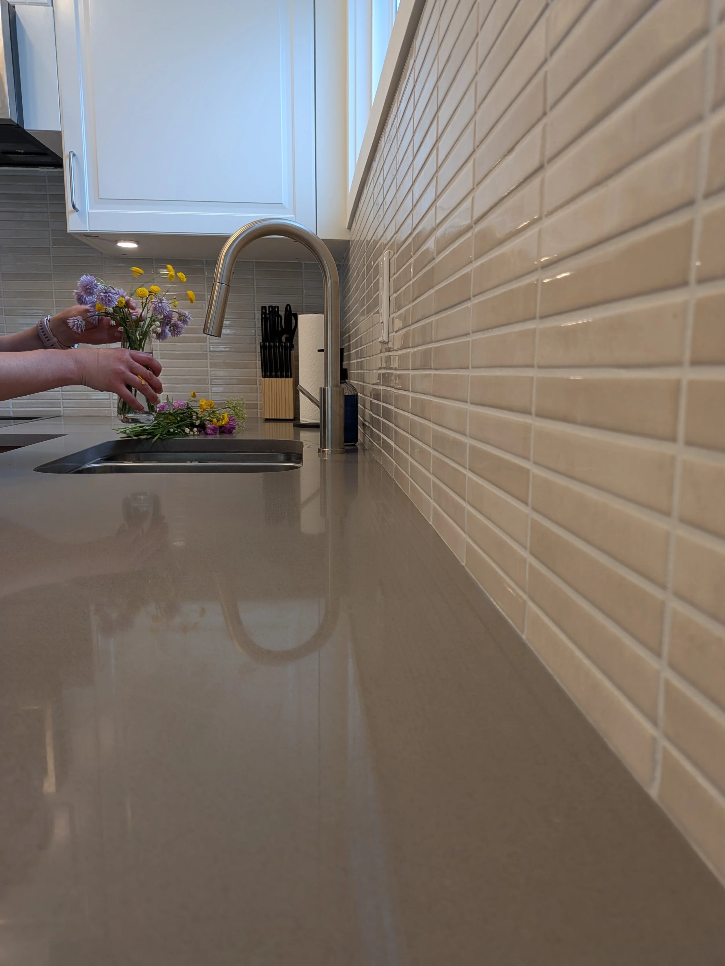 A kitchen sink with a person holding a small bouquet of purple and yellow flowers over it. The countertop is smooth, and there is a beige tiled backsplash. In the background, white kitchen cabinets and a window are visible.