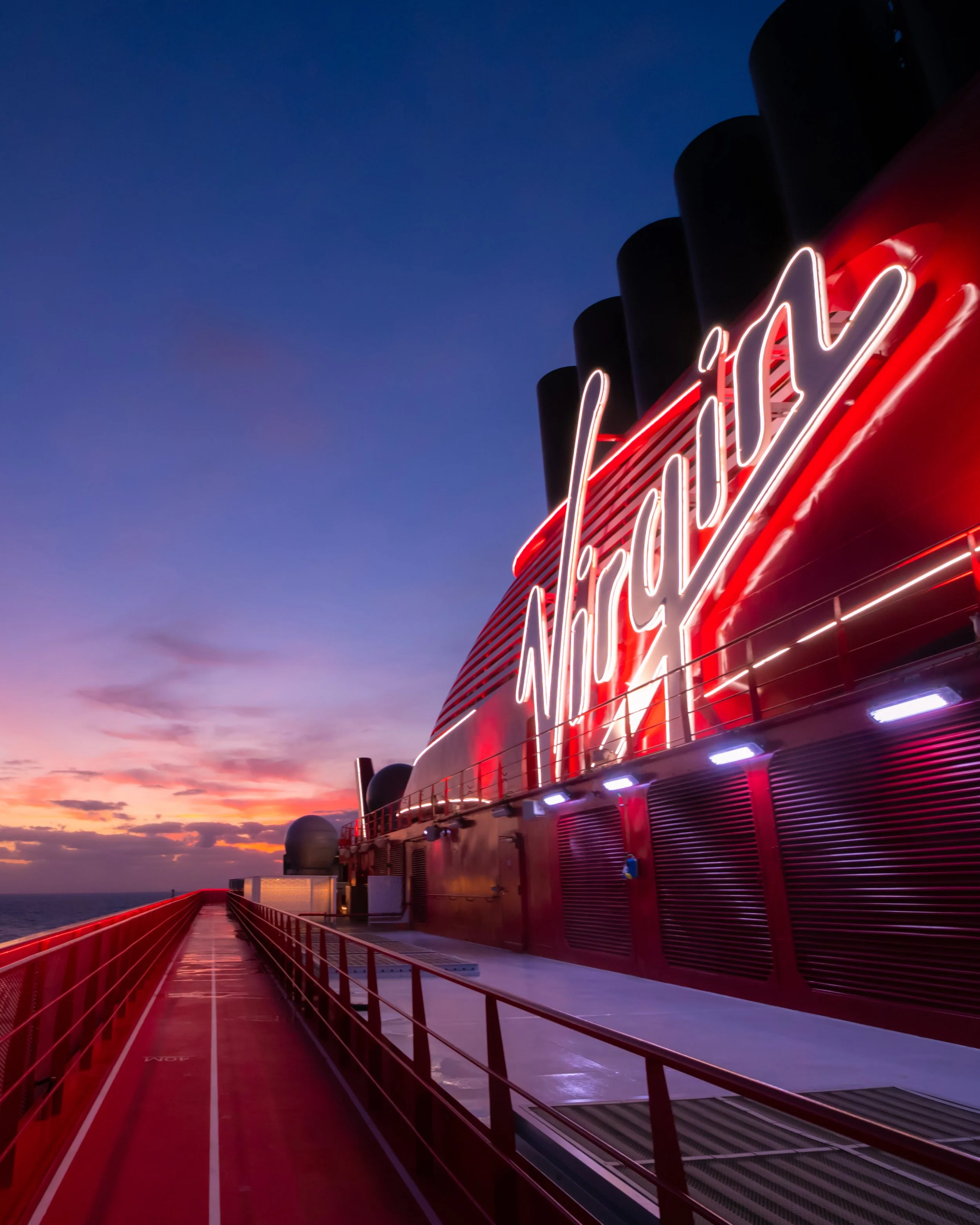 View of the deck of a cruise ship at sunset with a red neon sign and a red railing, featuring a modern building structure and an ocean horizon in the background.