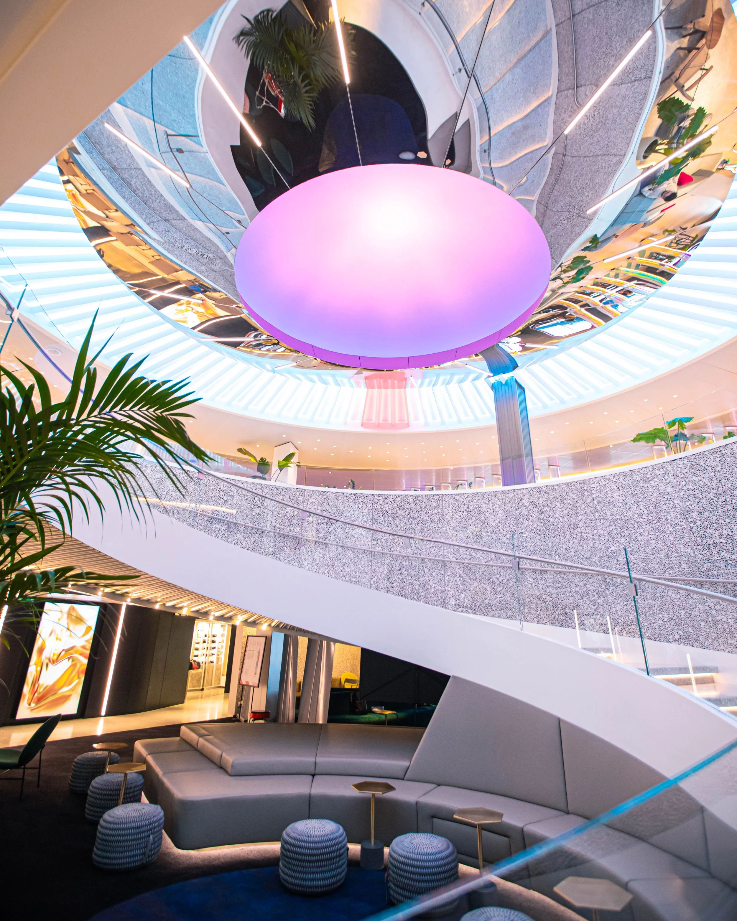 Virgin Voyages roundabout with a curved balcony, illuminated with soft LED lights. In the center, there's a circular purple-lit ceiling feature, plants, and sleek benches and stools for seating.