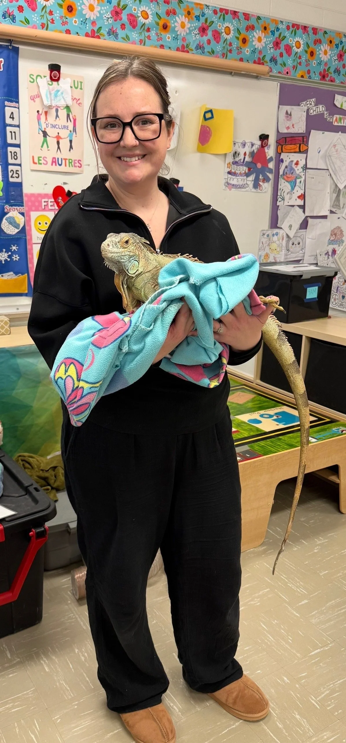 A woman wearing glasses and a black jacket holding a large iguana wrapped in a colorful towel inside a classroom. The classroom is decorated with educational posters, drawings, and colorful borders.