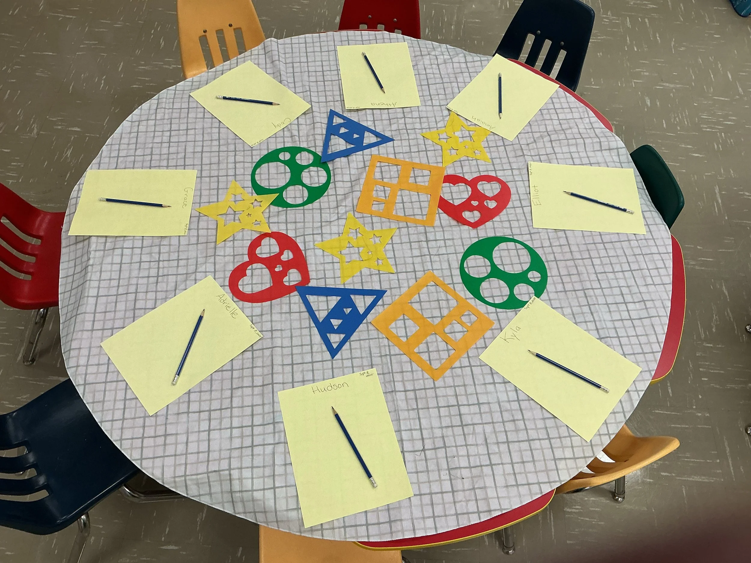 A round table covered with a checkered tablecloth. There are six yellow note cards with names and pens on top. In the center, there are colorful paper cutouts of various shapes, including stars, hearts, circles, squares, triangles, and a gear. The background shows chairs around the table and a patterned floor.