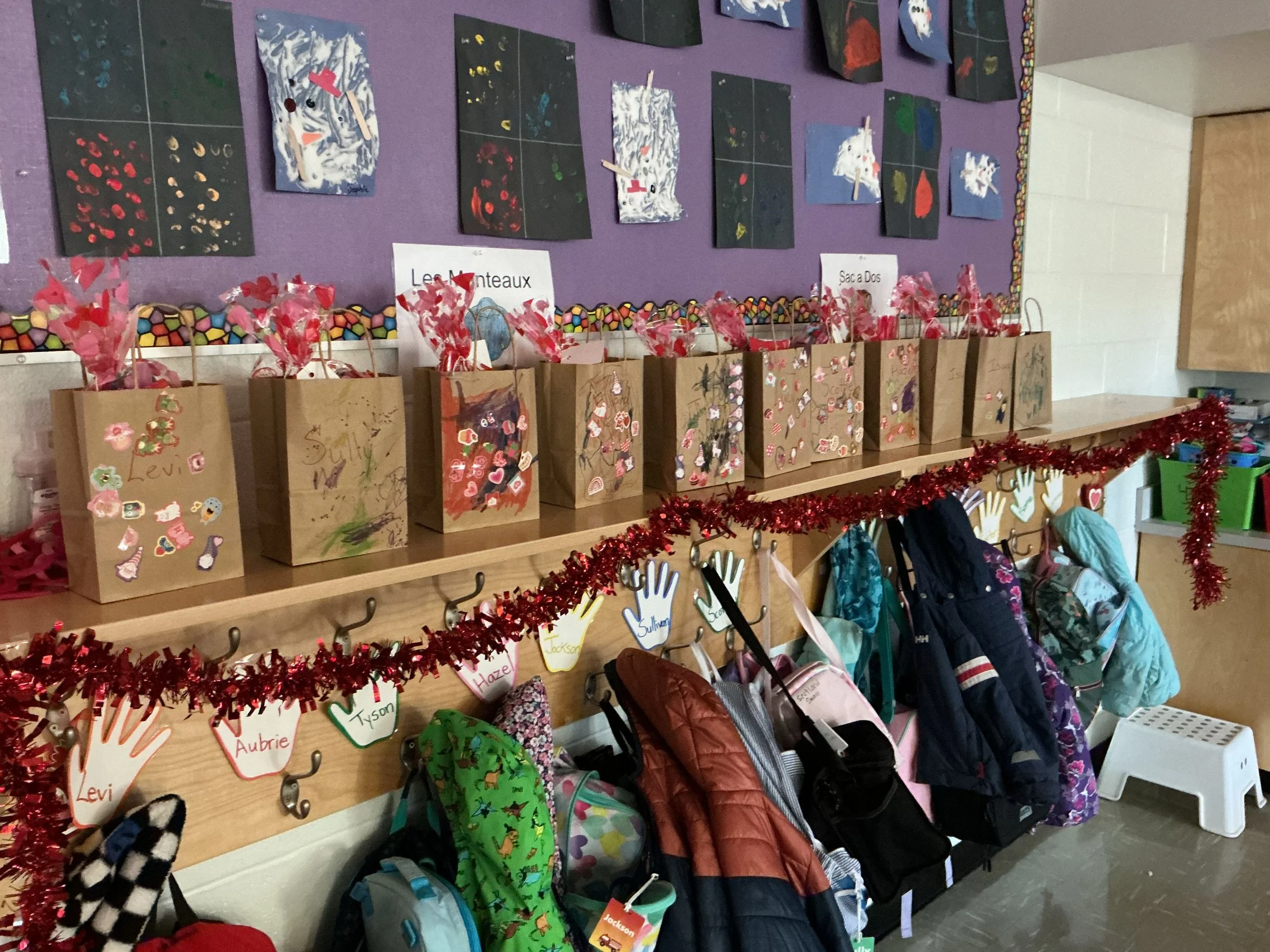 A classroom display with black and purple walls featuring student artwork and decorated paper bags with handwritten labels and children's names, backpacks hanging below, and red tinsel trim.