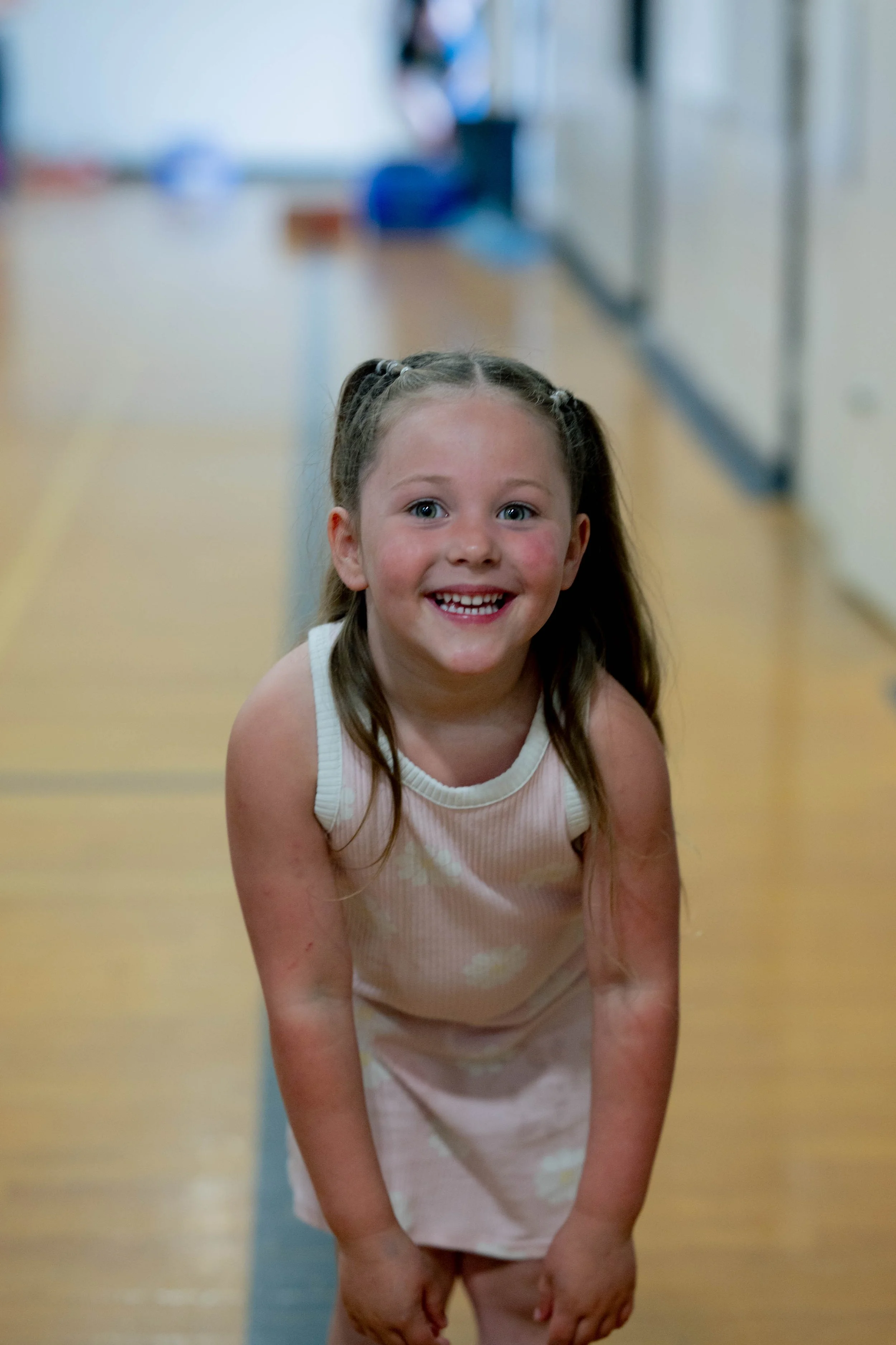 A young girl with brown hair styled in pigtails, smiling, kneeling on the floor in a school hallway, wearing a pink dress with white polka dots.