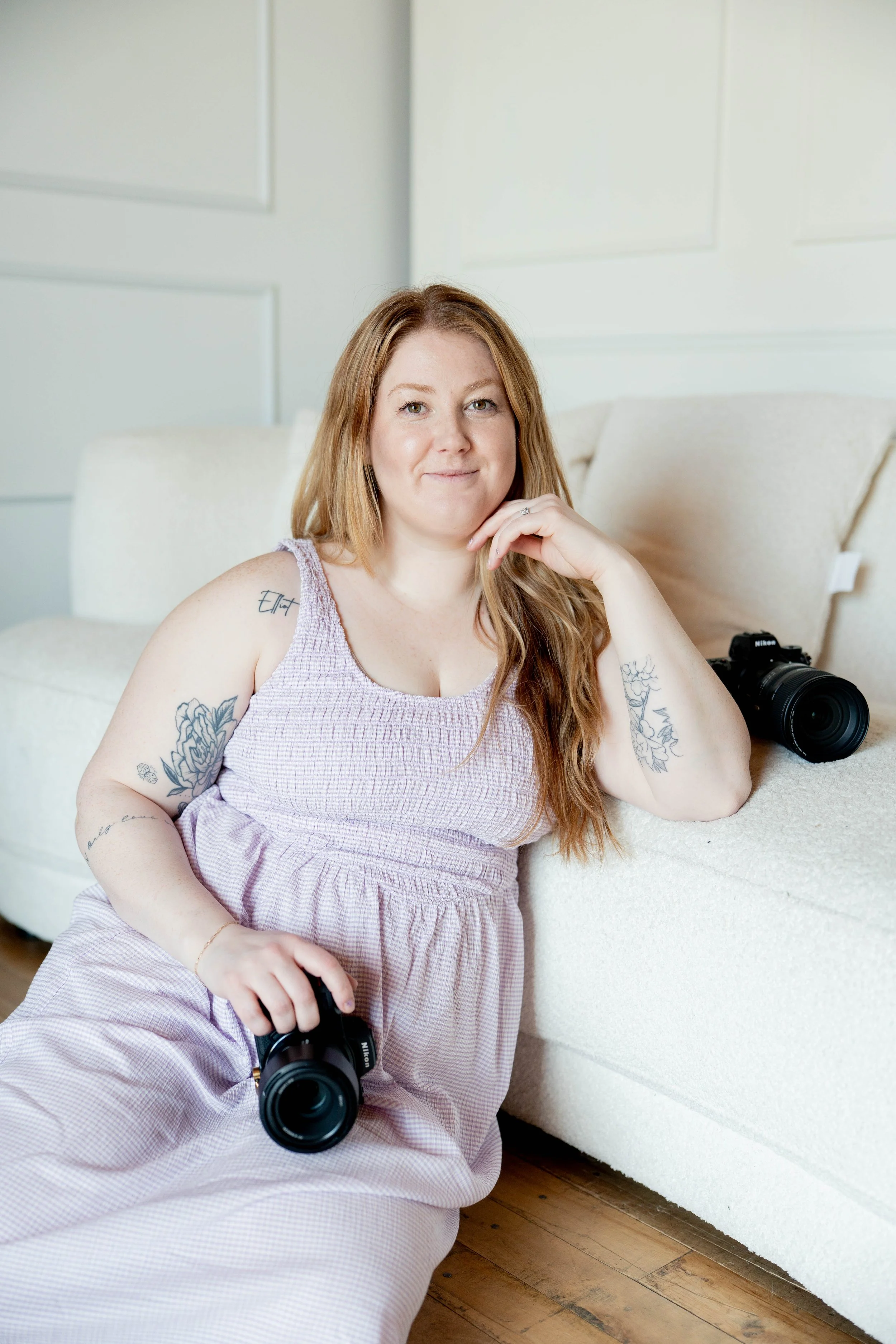 A woman with red hair and tattoos sitting on the floor, holding a camera in her hand, in a room with a white couch and walls, and a Nikon camera on the couch.