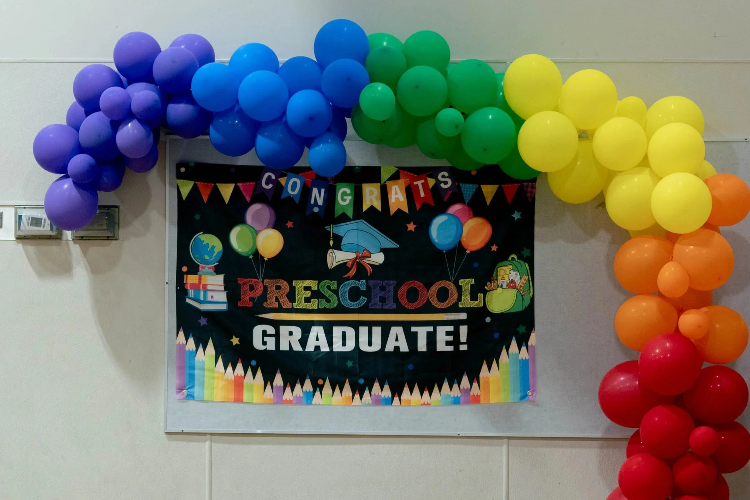 Colorful balloons arranged around a graduation celebration banner that reads 'Congratulations Preschool Graduate!' with educational and celebratory symbols.