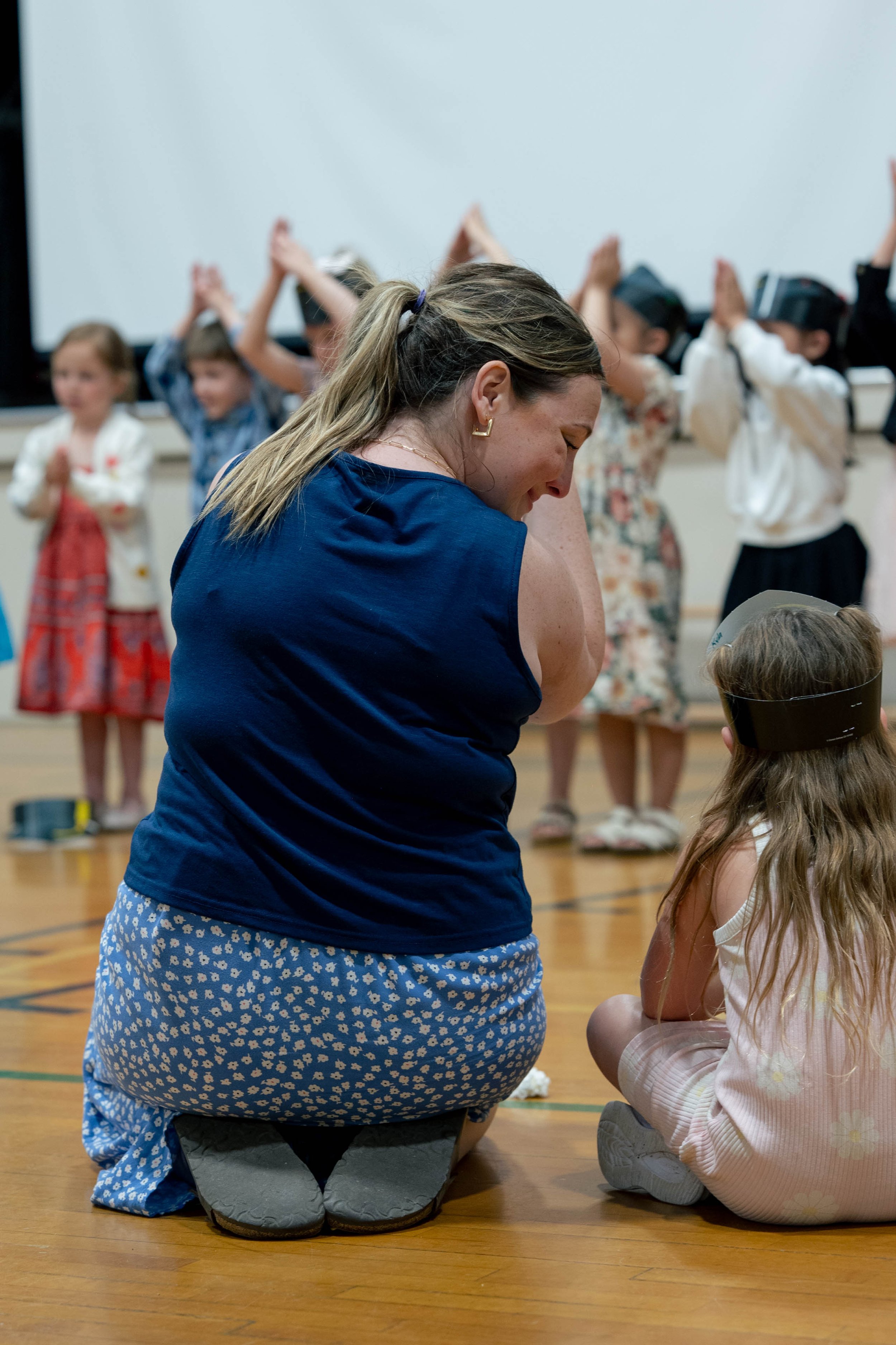 A woman kneeling on a gym floor with children in the background, some clapping and some wearing virtual reality headsets, in a classroom or indoor activity setting.