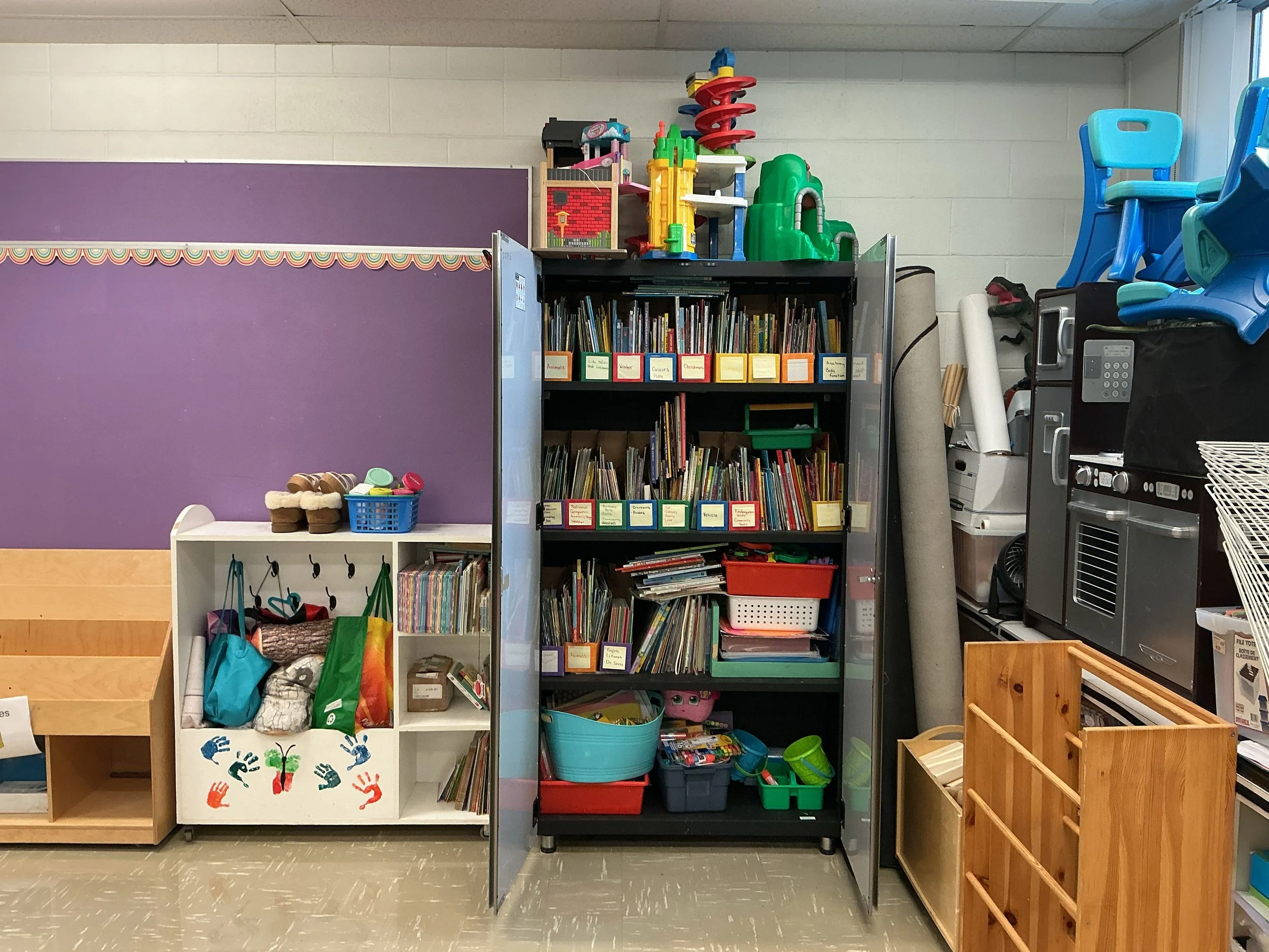 A classroom or library shelves filled with books, toys, and educational materials, with a purple wall and a window on the right side.