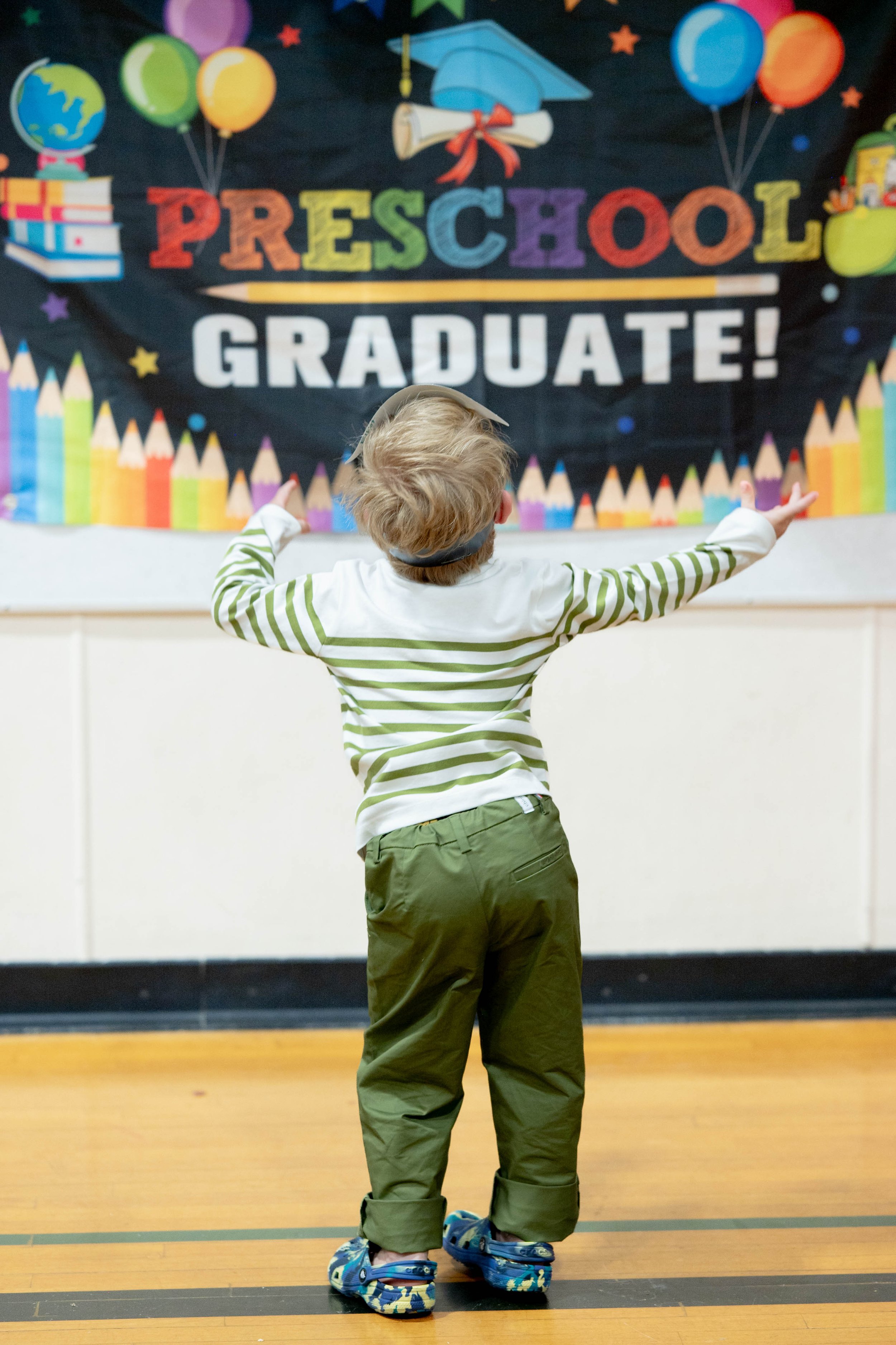 Child wearing striped shirt and green pants, front facing a diploma and graduation cap backdrop, with arms outstretched.