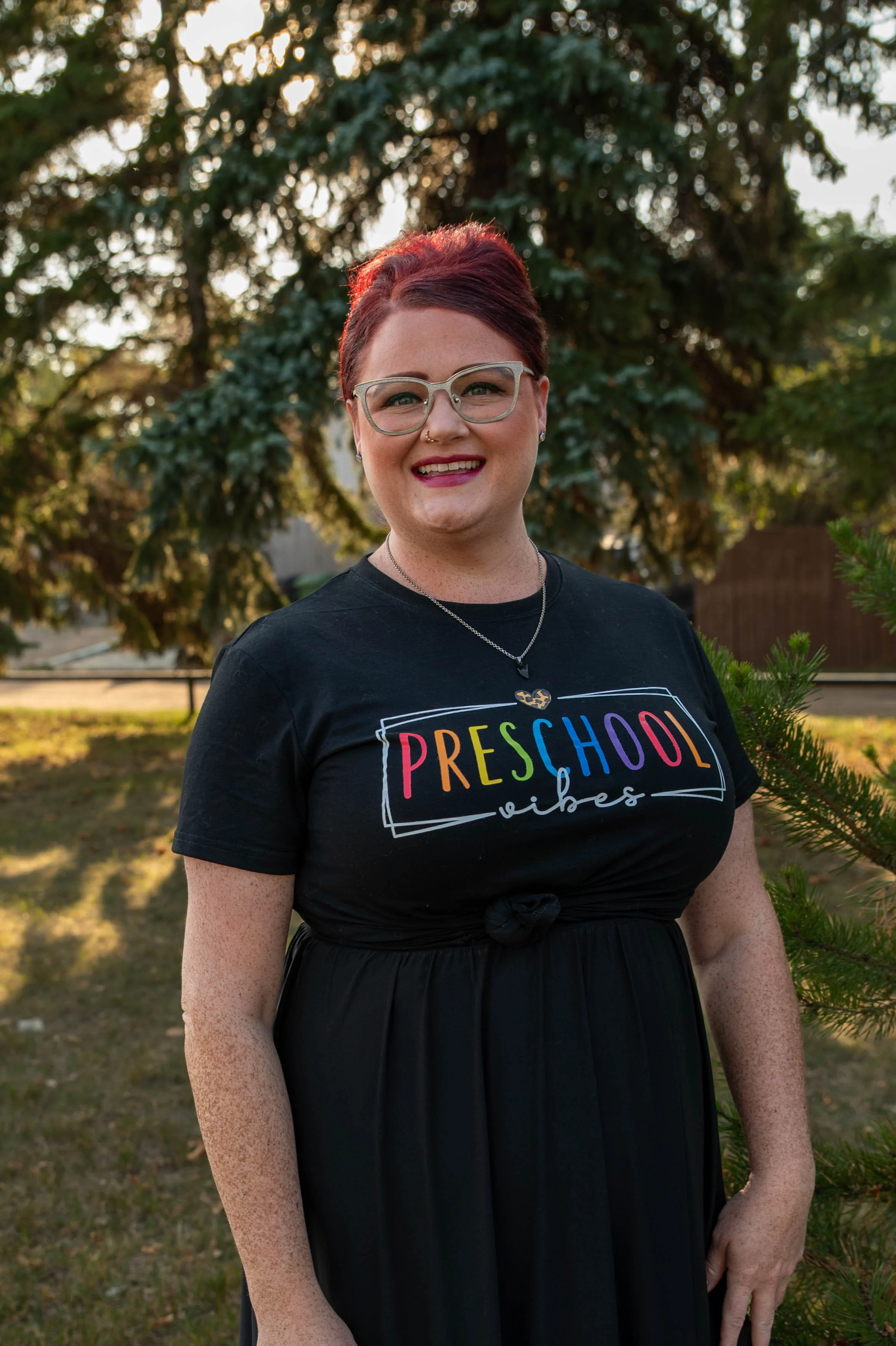 A woman with short red hair, glasses, and a nose piercing standing outdoors in front of a large tree, wearing a black T-shirt with rainbow-colored text that reads 'Preschool Vibes'.