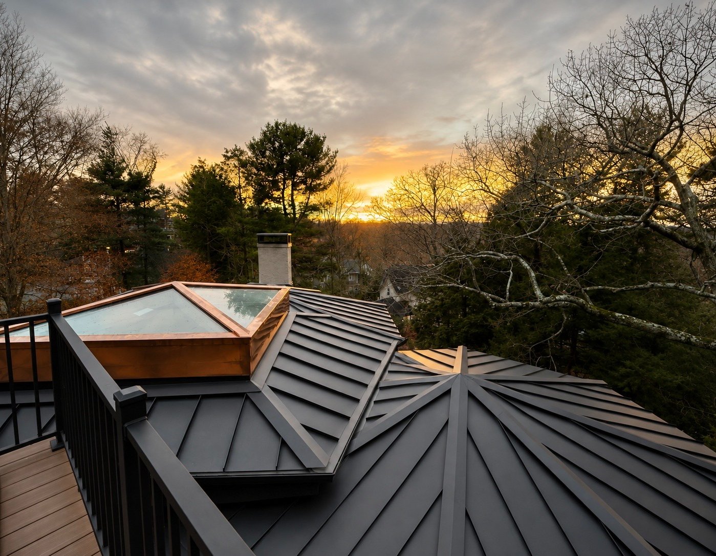 Sunset view from a rooftop featuring a skylight, metal roofing, bare trees, and a chimney, with a partly cloudy sky.