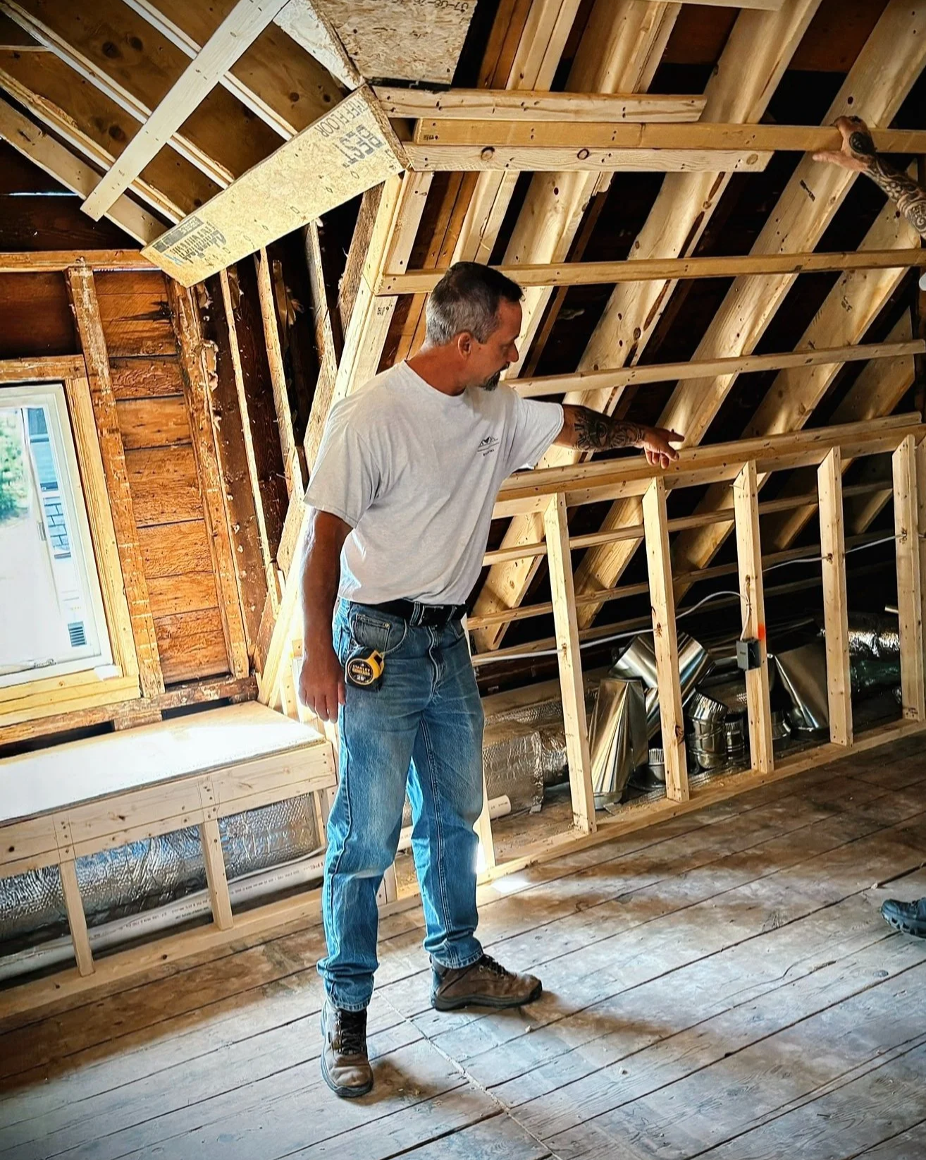 A man in a white t-shirt and jeans is standing inside a partially constructed room with a sloped ceiling. He is pointing at the wooden framing of the ceiling and wall, indicating construction or inspection of the unfinished space. There are tools and construction materials visible in the background.