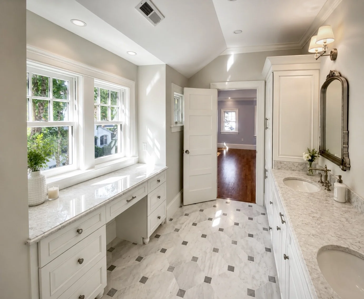 Bright bathroom with white cabinetry, marble countertops, and double sinks. Windows let in natural light, and a door leads to a room with hardwood floors.
