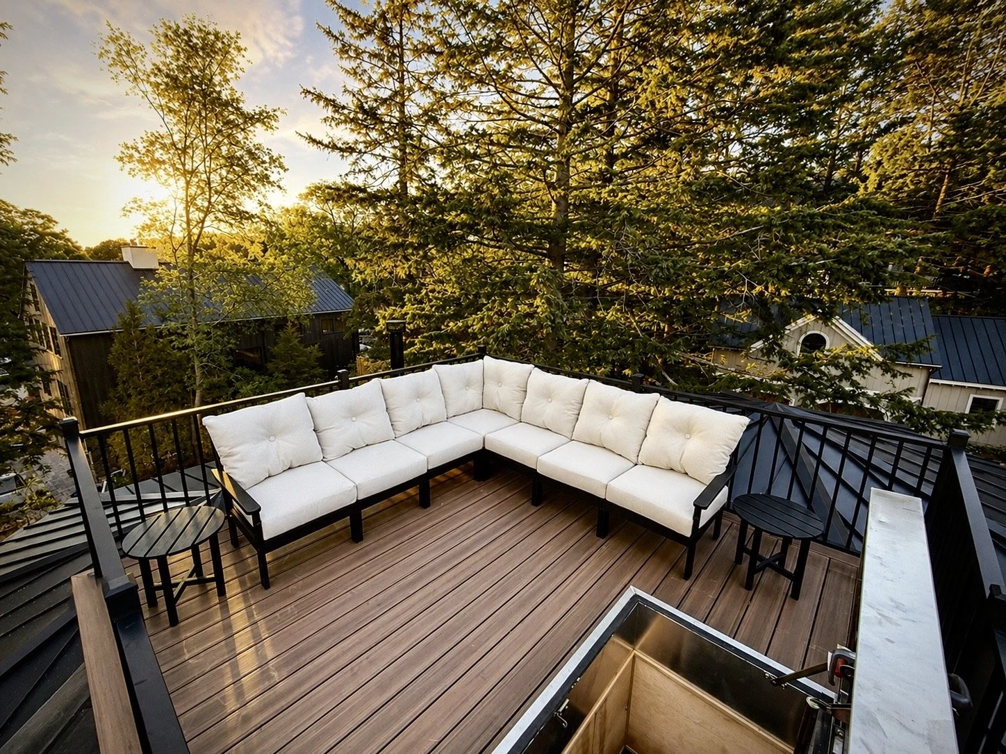 A rooftop deck with a black metal railing, brown wooden flooring, and outdoor white cushioned sectional sofa with throw pillows, overlooking trees and neighboring houses during sunset.