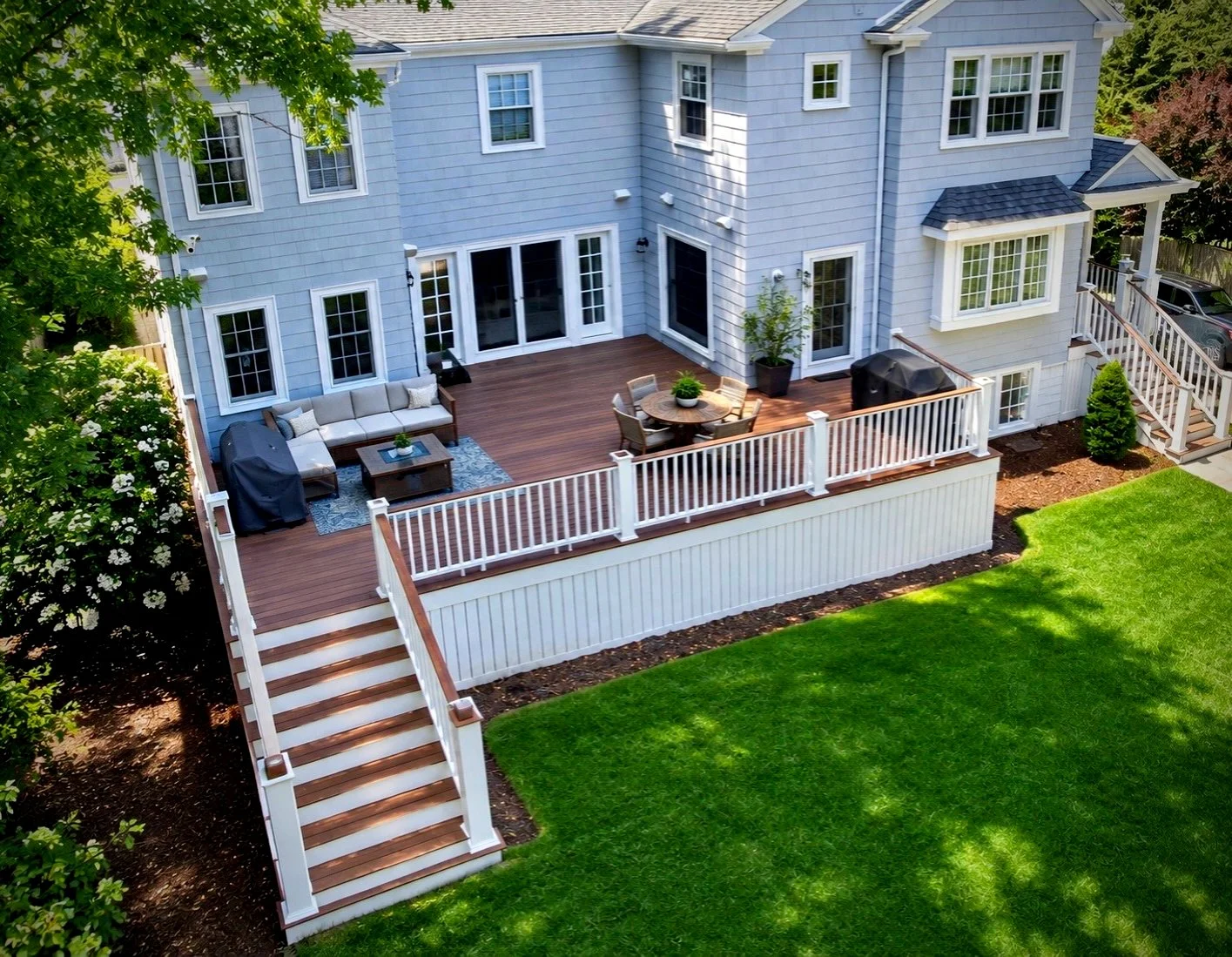 A spacious backyard deck attached to a light blue house with white trim. The deck has outdoor furniture including a sectional sofa, a round dining table with chairs, and a grill. It is surrounded by greenery, including a large tree, and bordered by w