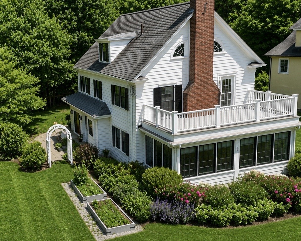 A white, multi-story house with black shutters, a brick chimney, and a spacious balcony. The house has a well-maintained garden with lush green grass and colorful flowering plants.