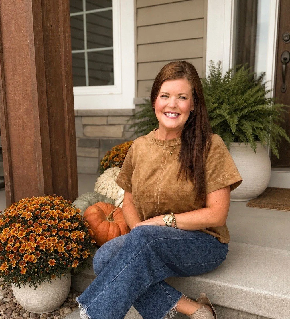 A woman sitting on front porch steps smiling, surrounded by pumpkins and fall flowers.