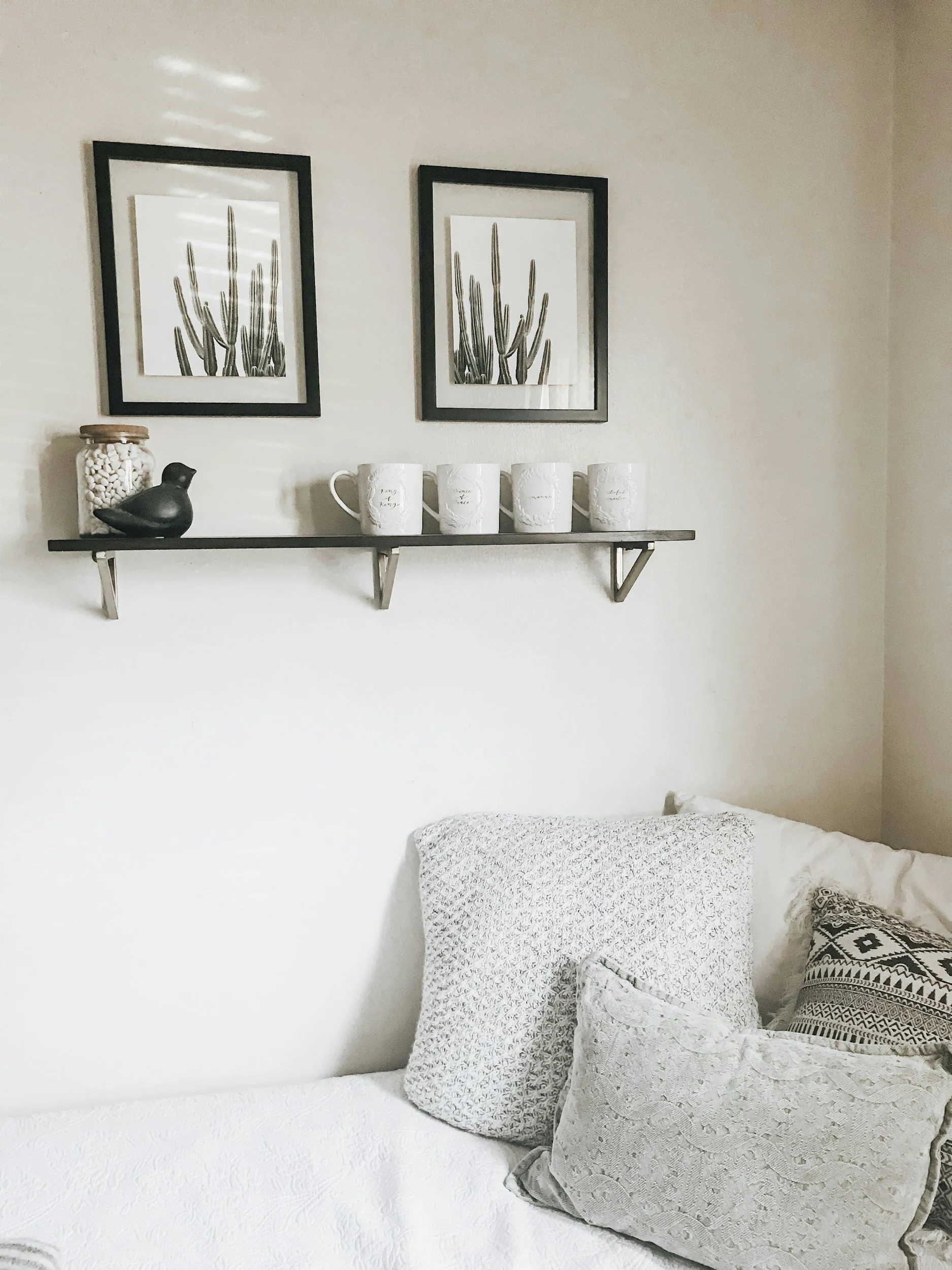 A bedroom corner with a white textured bedspread, multiple decorative pillows including one with black and white patterns, a wall-mounted black shelf with a black bird figurine, a jar of white candies, and four white mugs with scripted writing. Above the shelf are two black picture frames containing images of cacti.