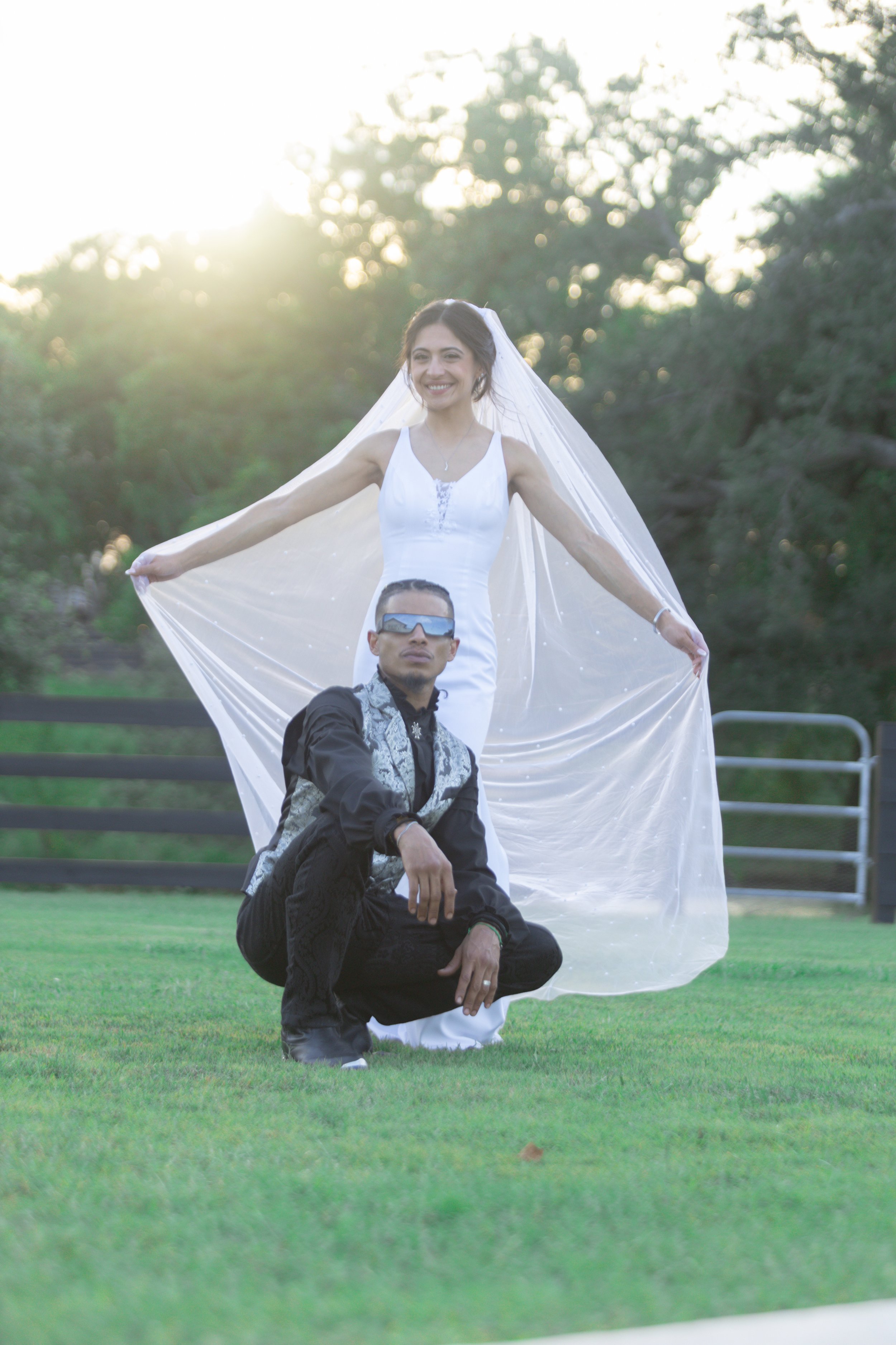 A bride in a white wedding dress with a veil standing outdoors while smiling, with a man in black clothing kneeling in front of her. The man is wearing sunglasses and has a serious expression. The background features trees and sunlight.