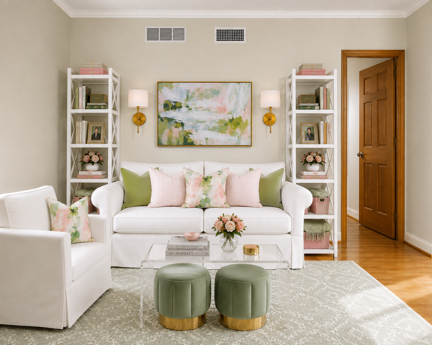 Bright living room with white sofa, pink and green pillows, two white bookshelves, abstract artwork, wall-mounted lamps, wooden door, glass coffee table with flowers, two green ottomans, beige patterned rug, wood floors, and beige walls.