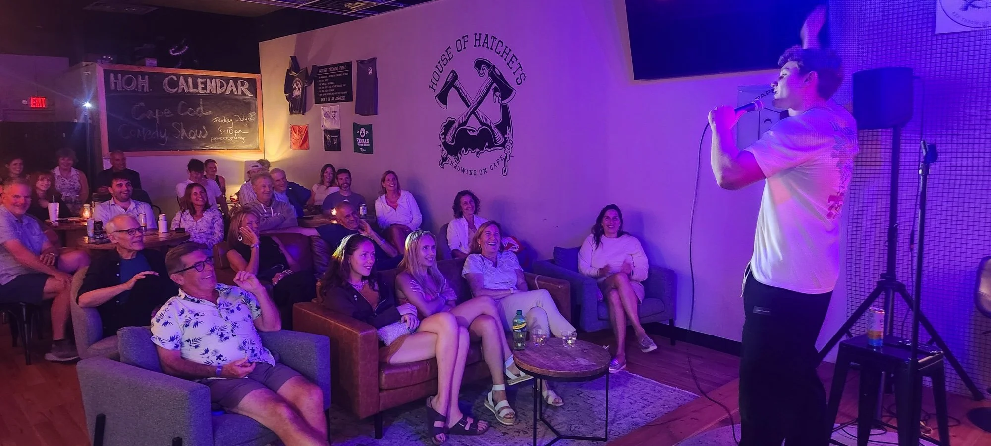 A comedy show in a bar with an audience laughing and a comedian on stage singing into a microphone, with posters and signs on the wall.