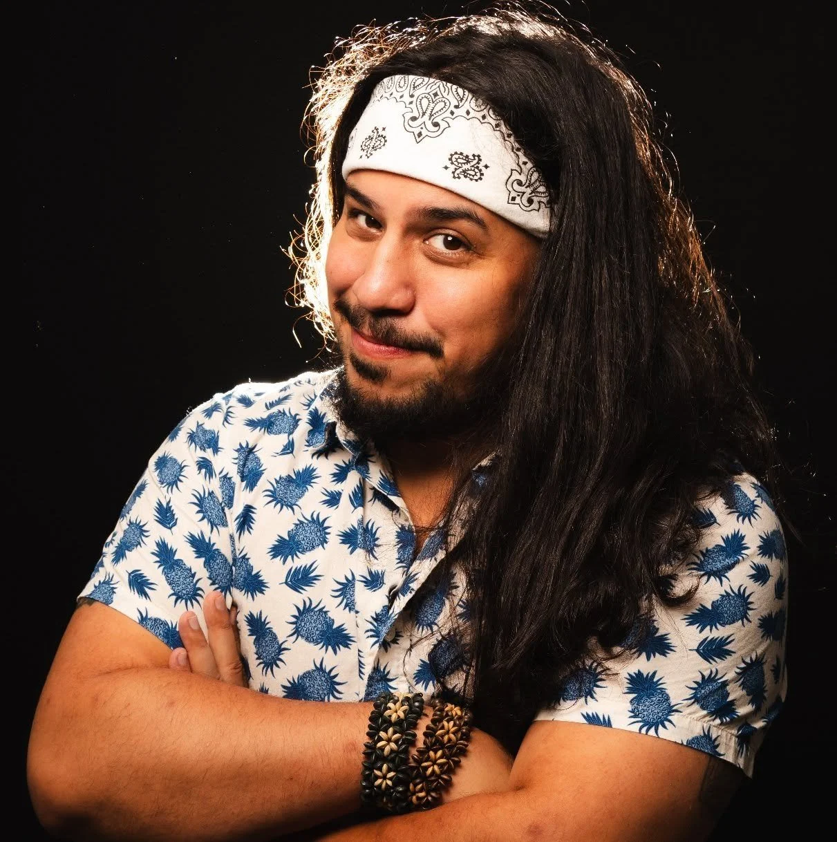 A man with long hair, a bandana, and a patterned shirt poses with a confident smile against a black background.
