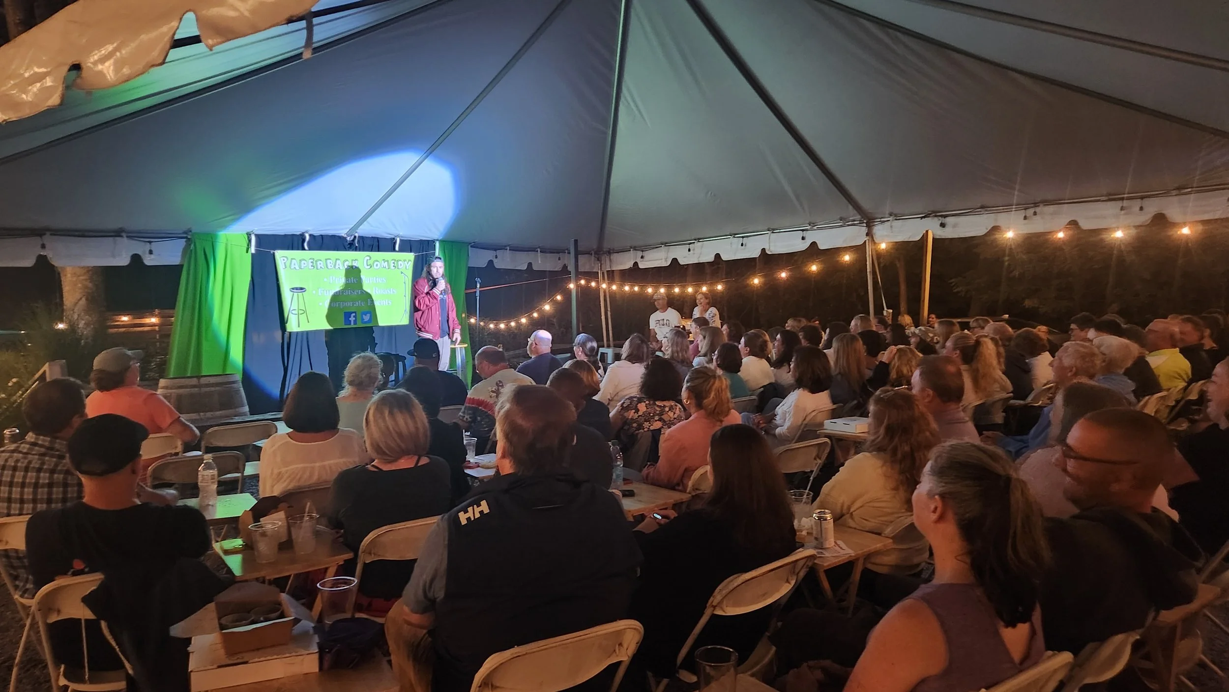 People attending a comedy show inside a large tent with string lights. A performer is on stage with a green curtain and a sign that reads "Paperback Comedy".