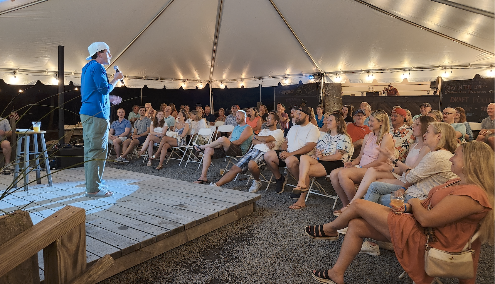 An outdoor comedy show under a large tent at night. A comedian in a blue shirt and baseball cap performs on a small wooden stage, holding a microphone. Audience members are seated on white folding chairs, laughing and enjoying the show. Some are holding drinks, and the setting is lively and relaxed.