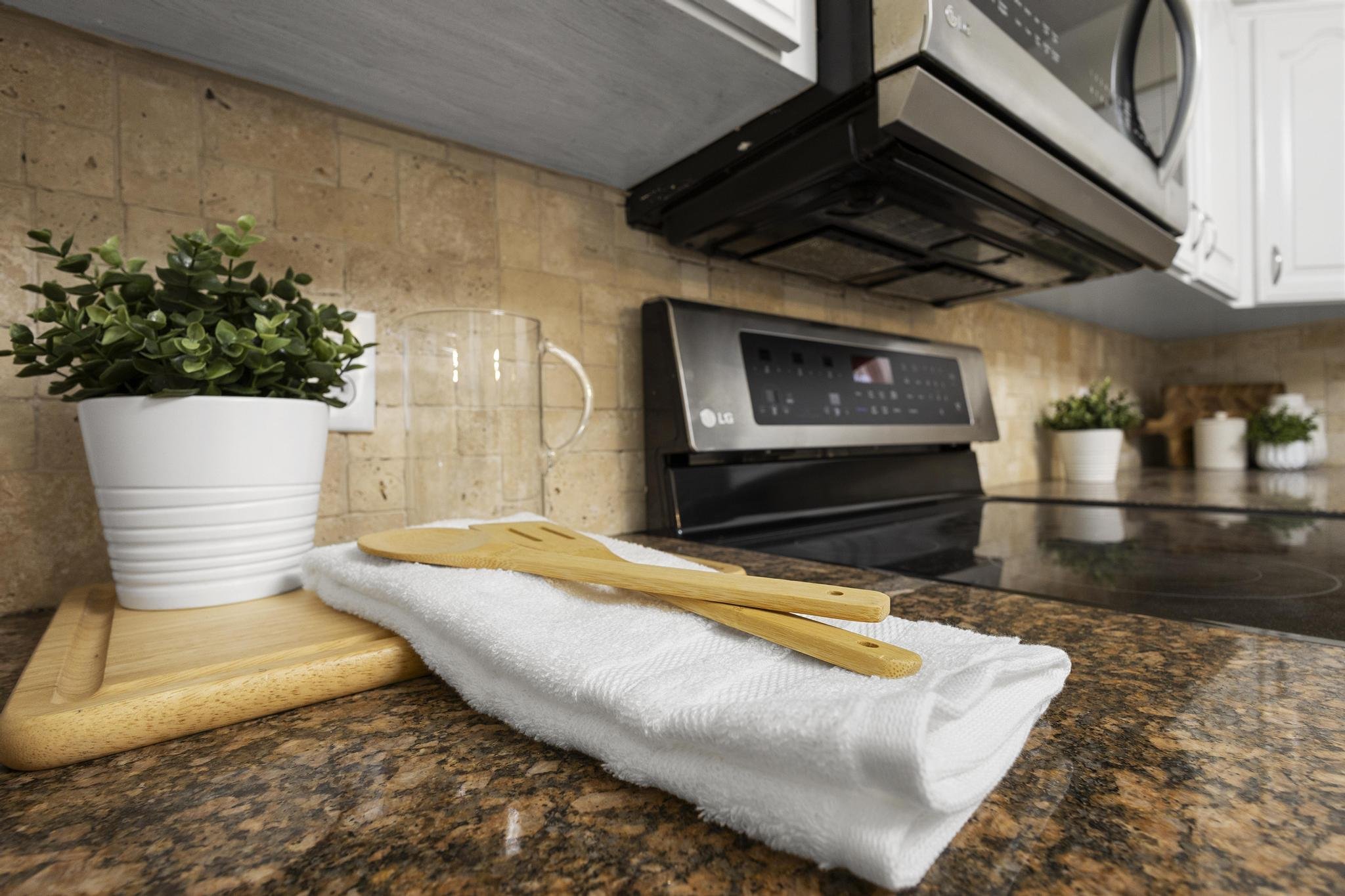 Kitchen countertop with potted plant, cutting board, towel, and stovetop. Cabinets and microwave in the background.