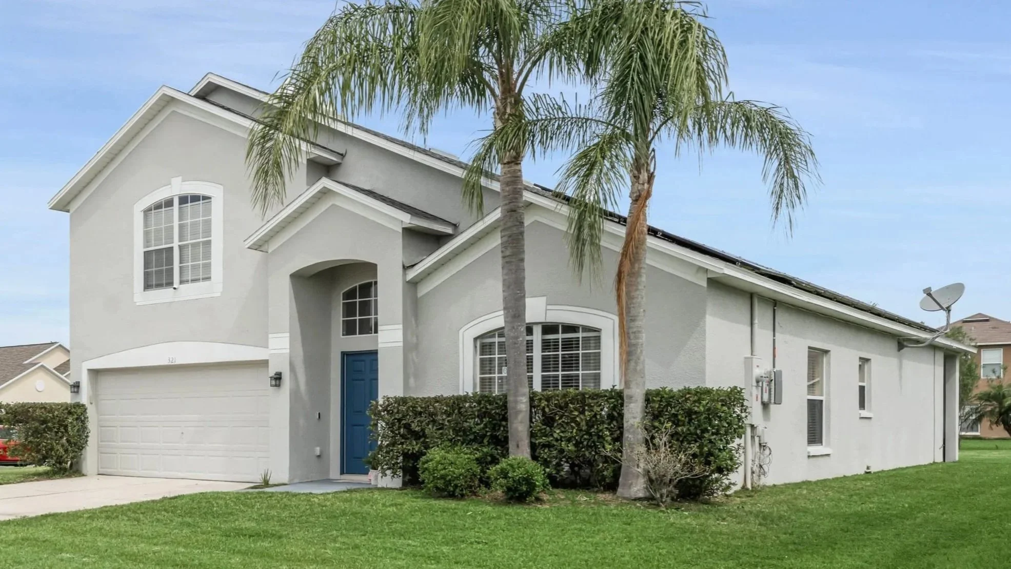 A modern two-story house with a white exterior, blue front door, and large windows. There are two tall palm trees in the front yard, along with neatly trimmed bushes and a well-maintained lawn. A satellite dish is mounted on the side of the house.