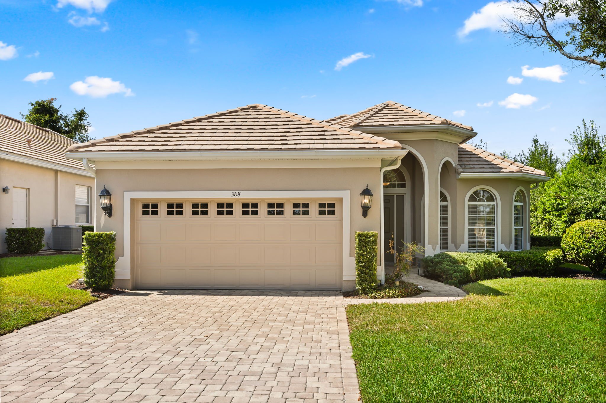 Front view of a single-story house with a beige garage door, white trim, and a small porch with a curved design. The house has a tiled roof and is surrounded by green grass, shrubs, and trees under a blue sky with a few fluffy clouds.