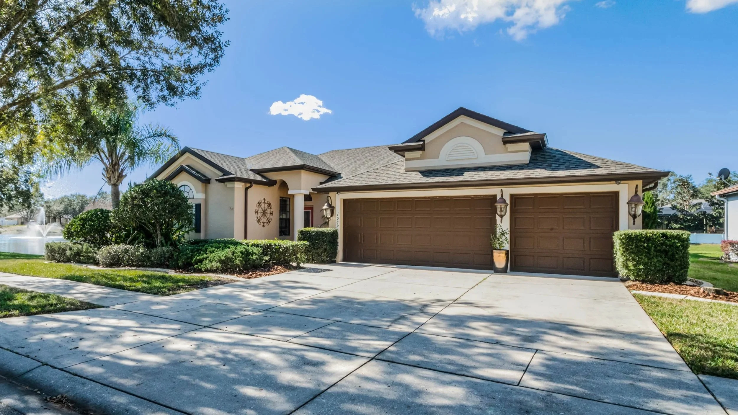 A house with a brown garage door, beige exterior walls, and a shingled roof. There are two outdoor lantern-style lights on either side of the garage. The front yard has green bushes, a small tree, and a fountain in the background. The sky is blue with a few clouds.