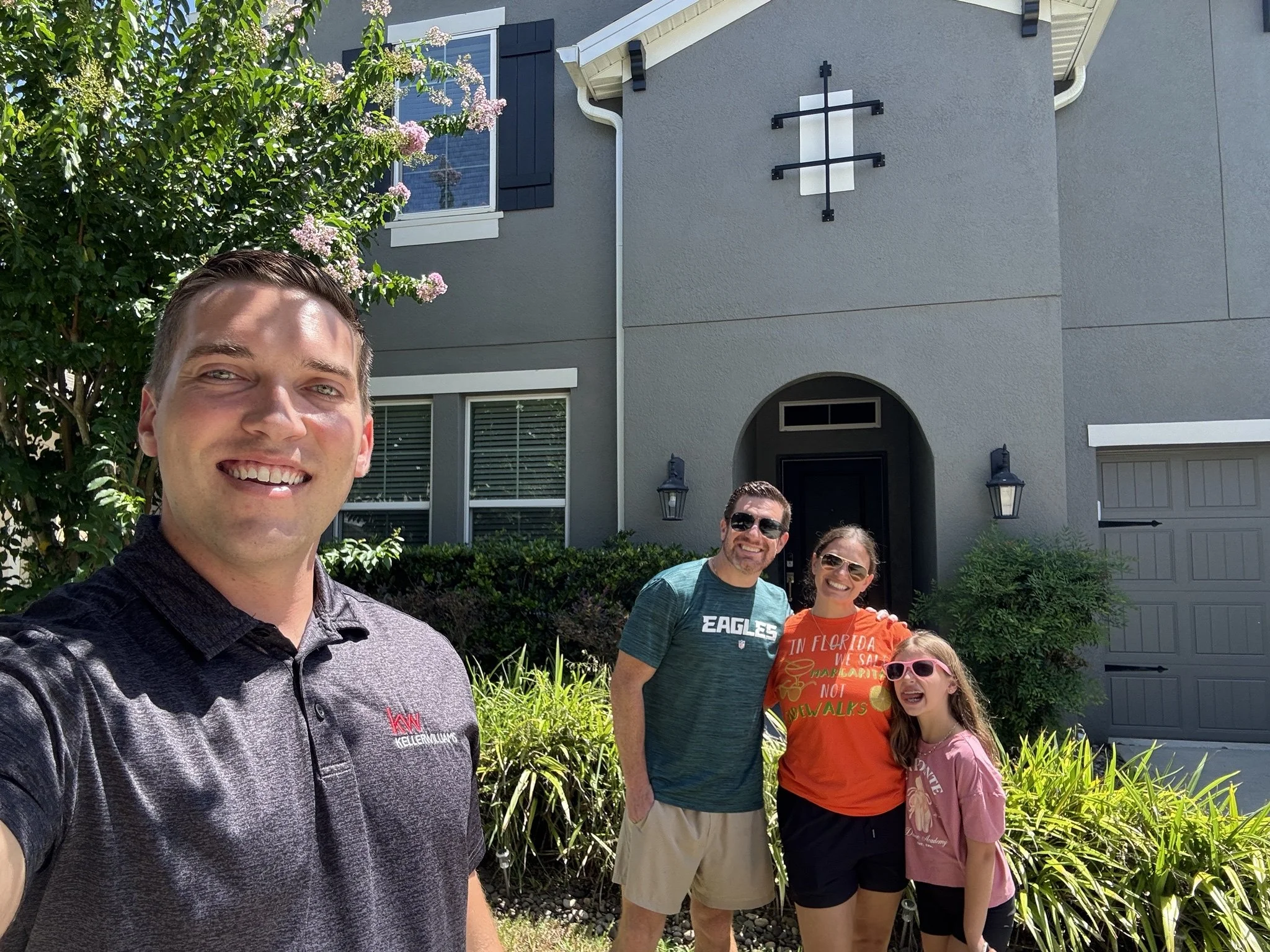 Group of four people standing outdoors in front of a gray house with black shutters, smiling at the camera, with greenery and flowering trees around.