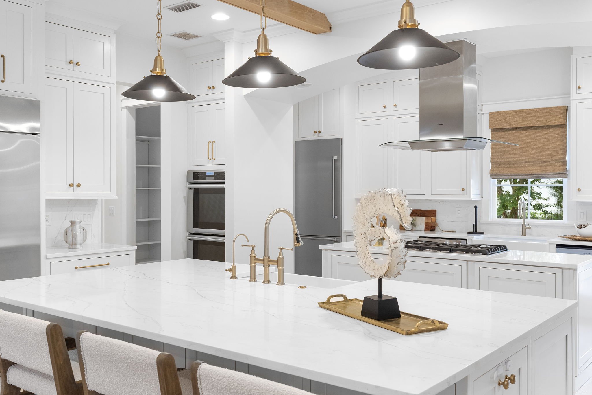 White kitchen with marble island, gold fixtures, black pendant lights, stainless steel appliances, and a window with a brown woven window shade.