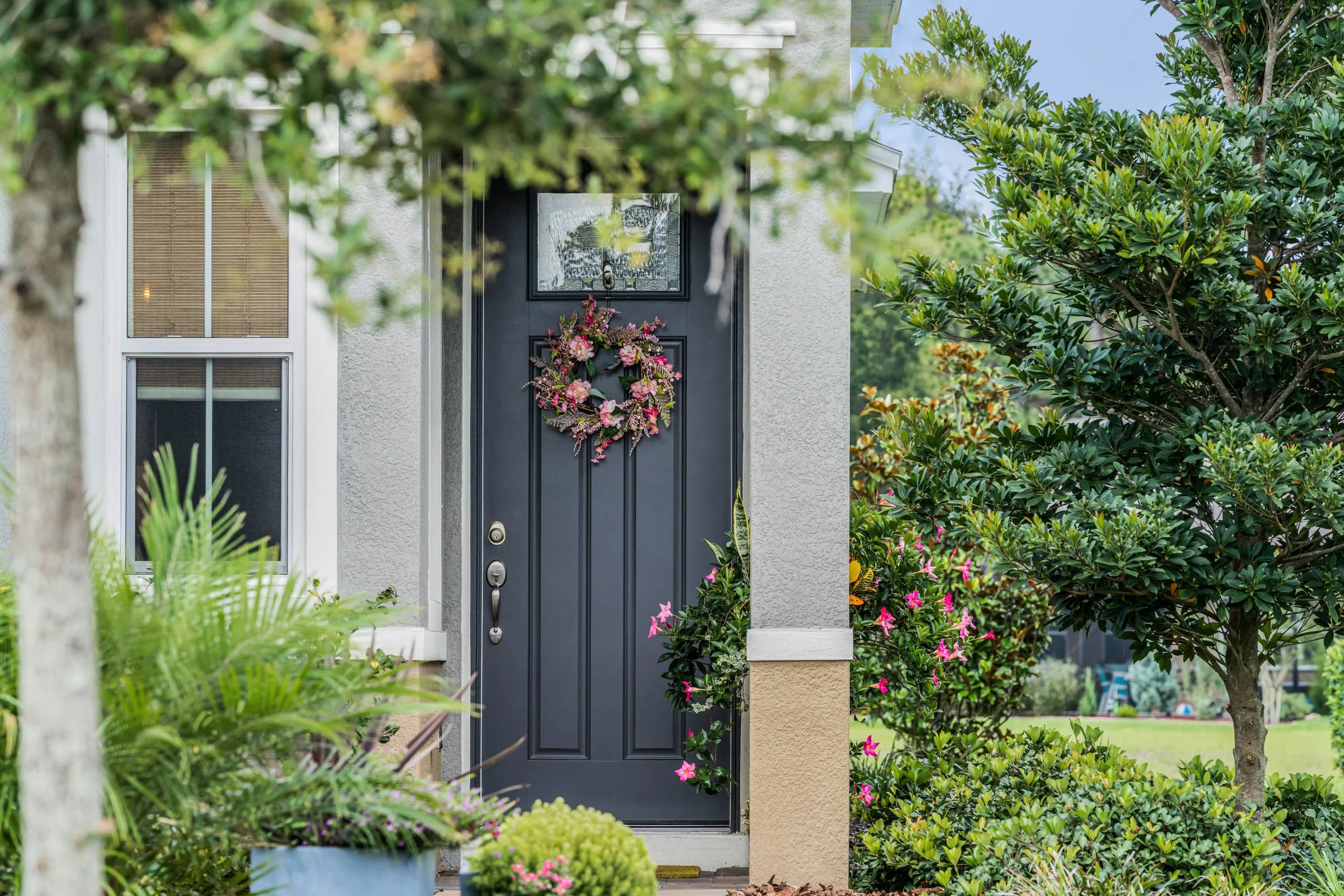 Front door decorated with a pink floral wreath, partially obscured by surrounding green bushes and trees.