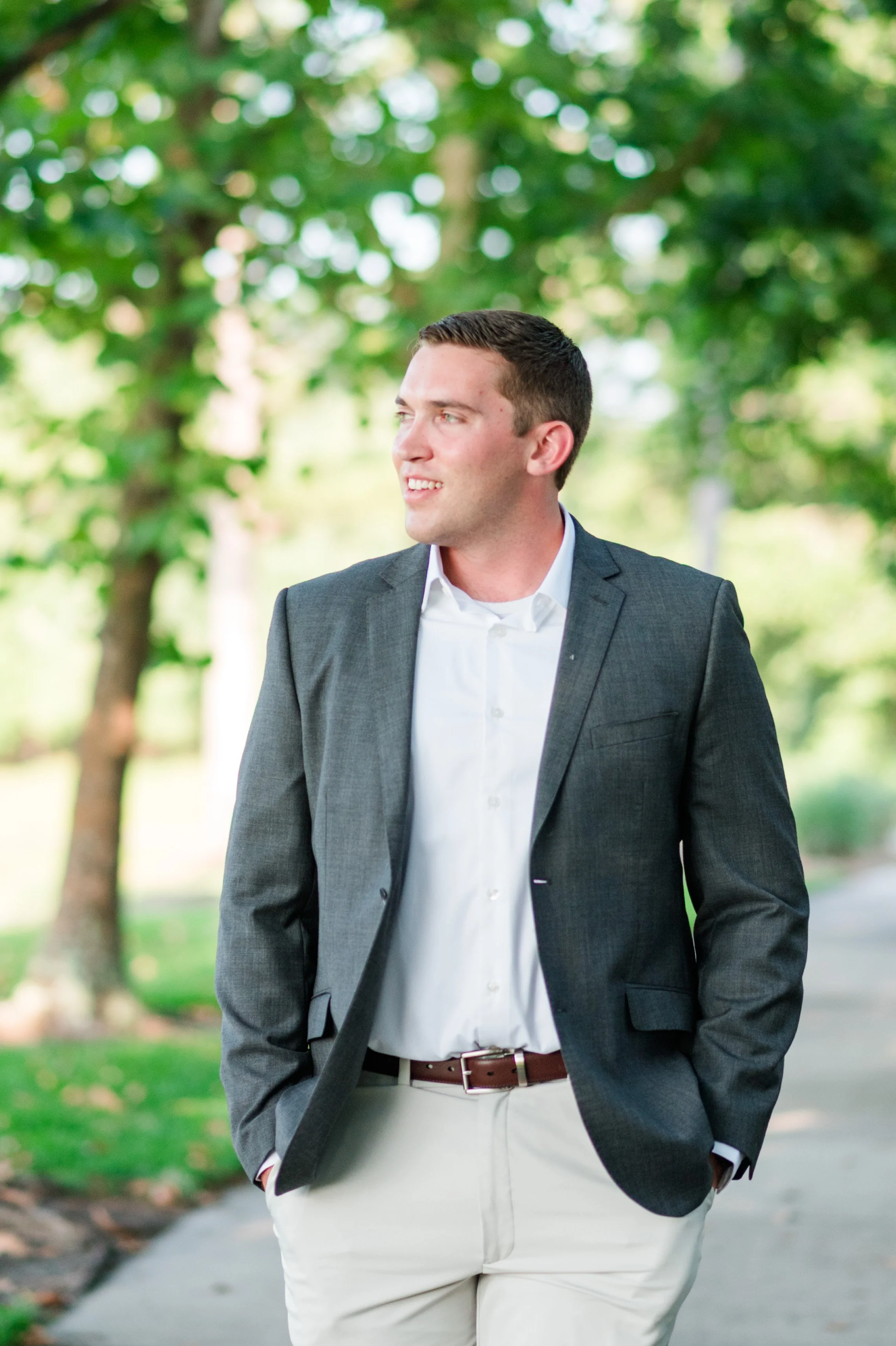 A man wearing a grey blazer, white shirt, and beige pants walking outdoors with trees in the background.
