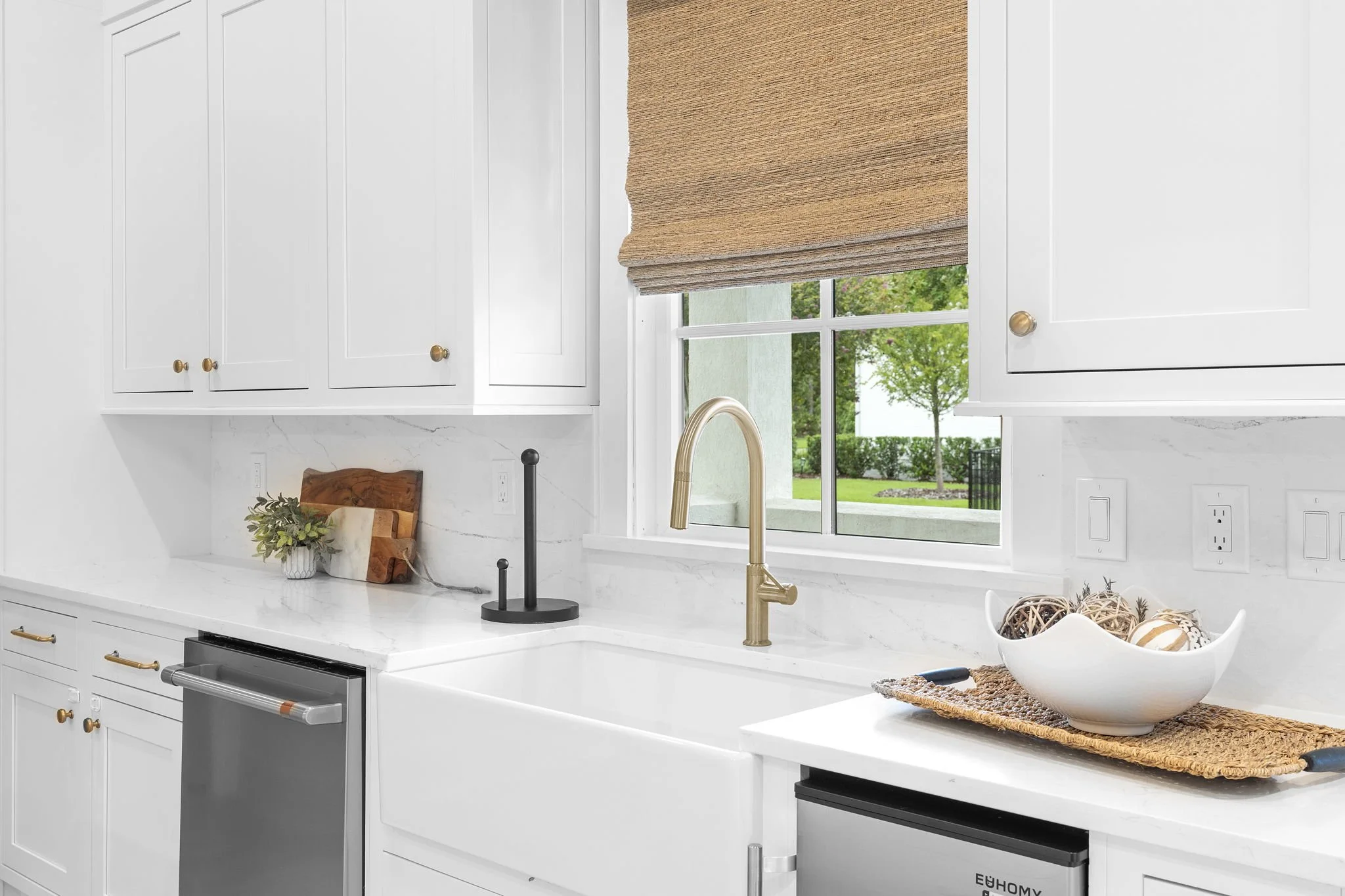 Kitchen with white cabinets, a window with a brown shade, a gold faucet, a sink, a cutting board, a plant, and a decorative bowl with balls.