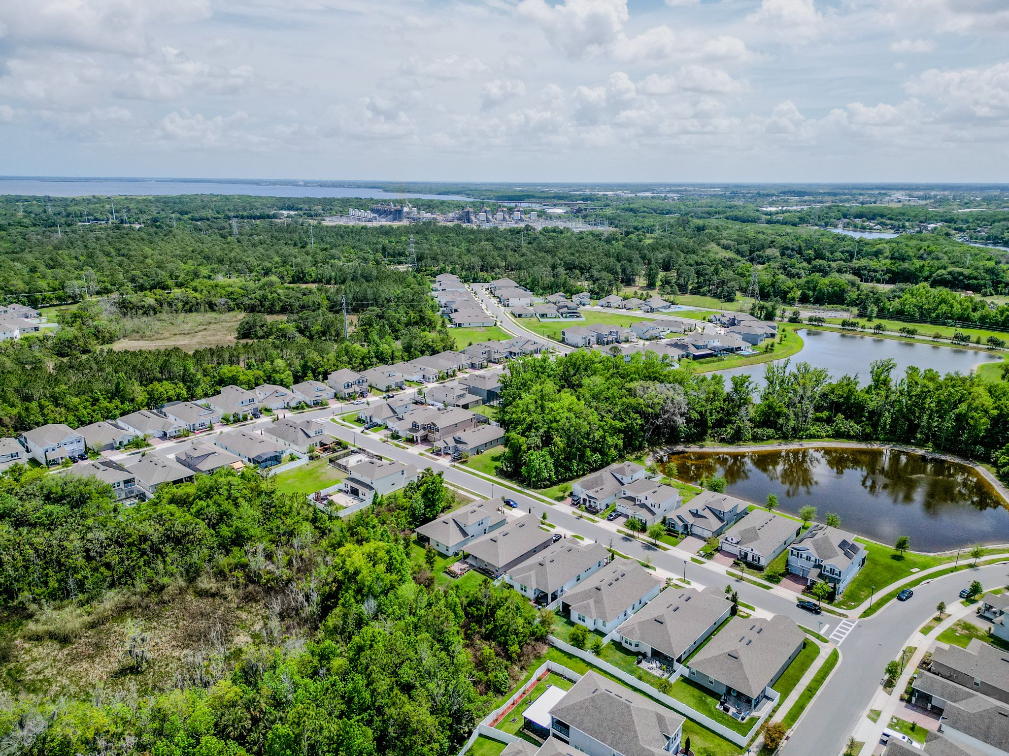 Aerial view of a suburban neighborhood with houses, a pond, trees, and a wooded area, with a large industrial complex and a river in the distance under a partly cloudy sky.