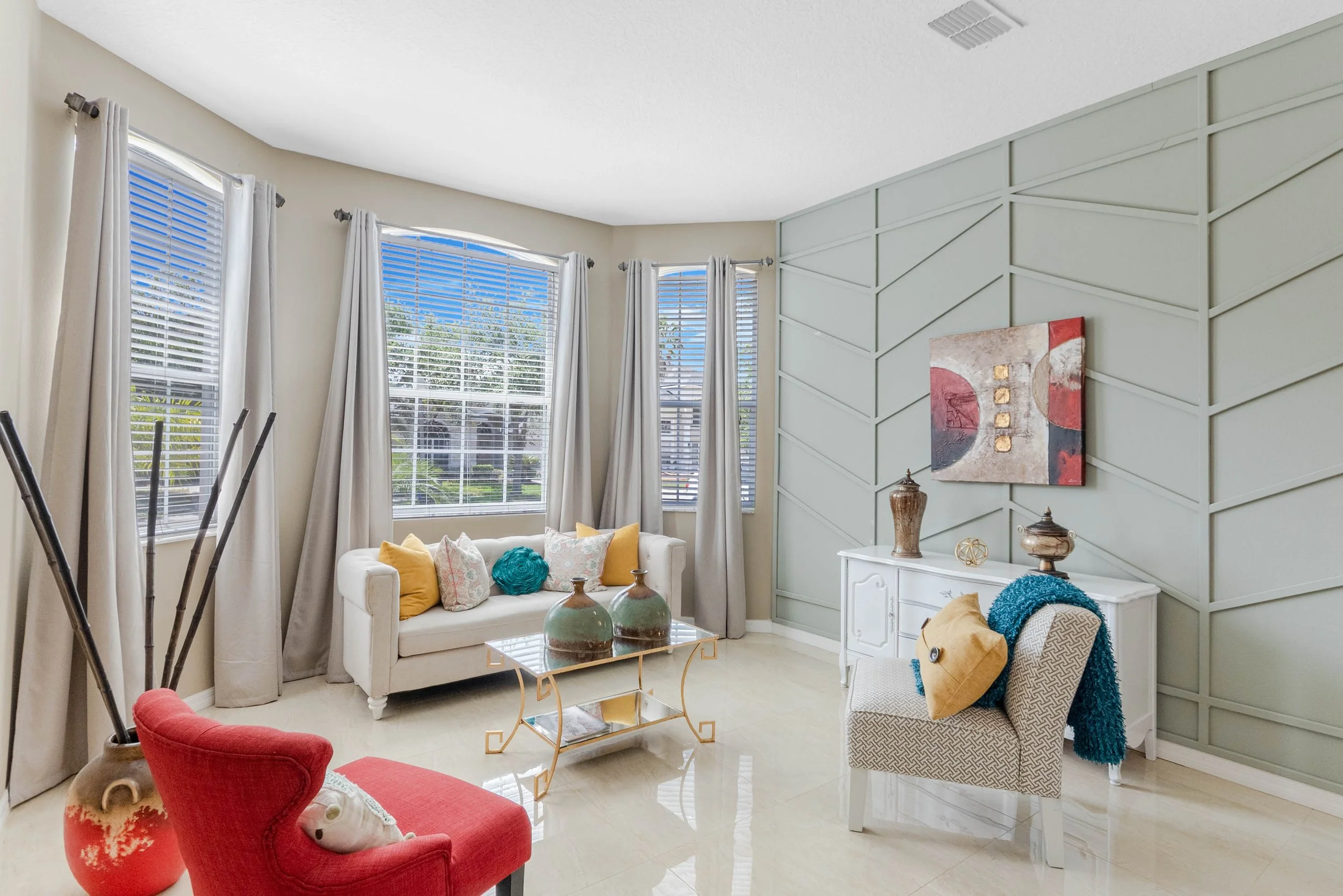 A bright living room with large windows, white curtains, a white sofa with colorful pillows, a gold coffee table, a red armchair, and decorative vases and artwork on the walls.